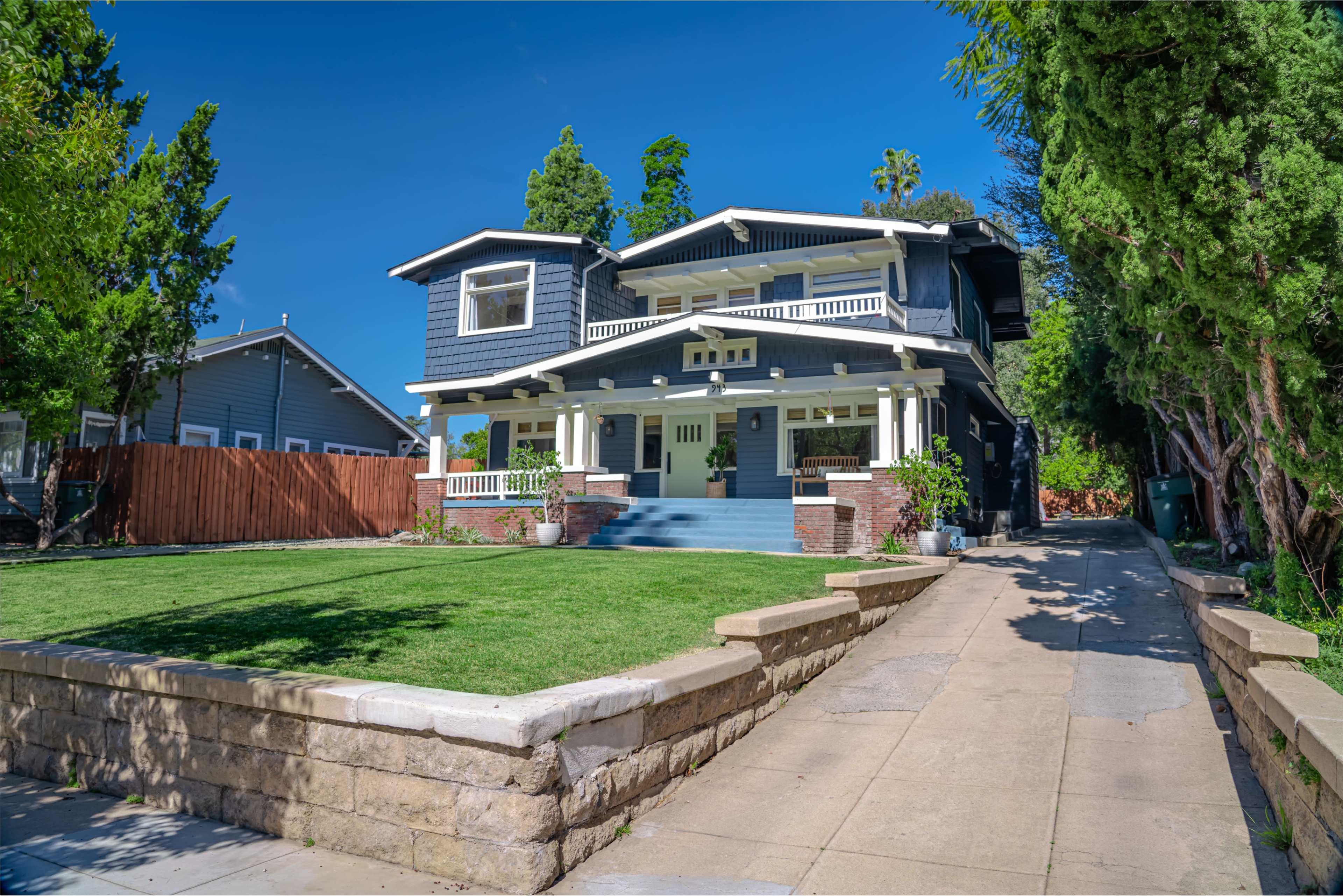 A large, two-story blue house with a welcoming front porch and neatly landscaped yard is situated on a sloped driveway.