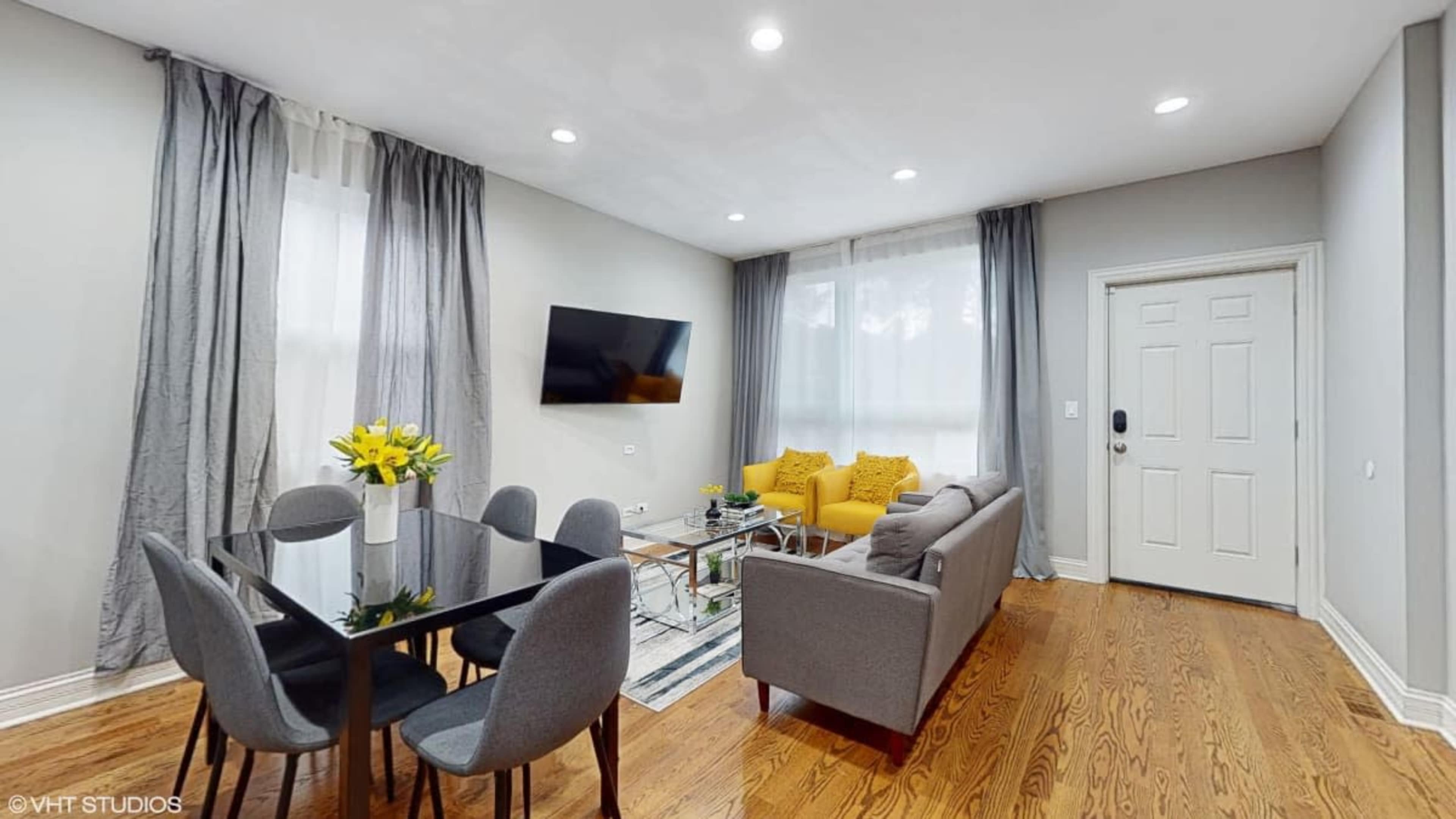 The image shows a modern living room featuring a gray sofa, a glass dining table with black chairs, and yellow accent pillows on the couch.