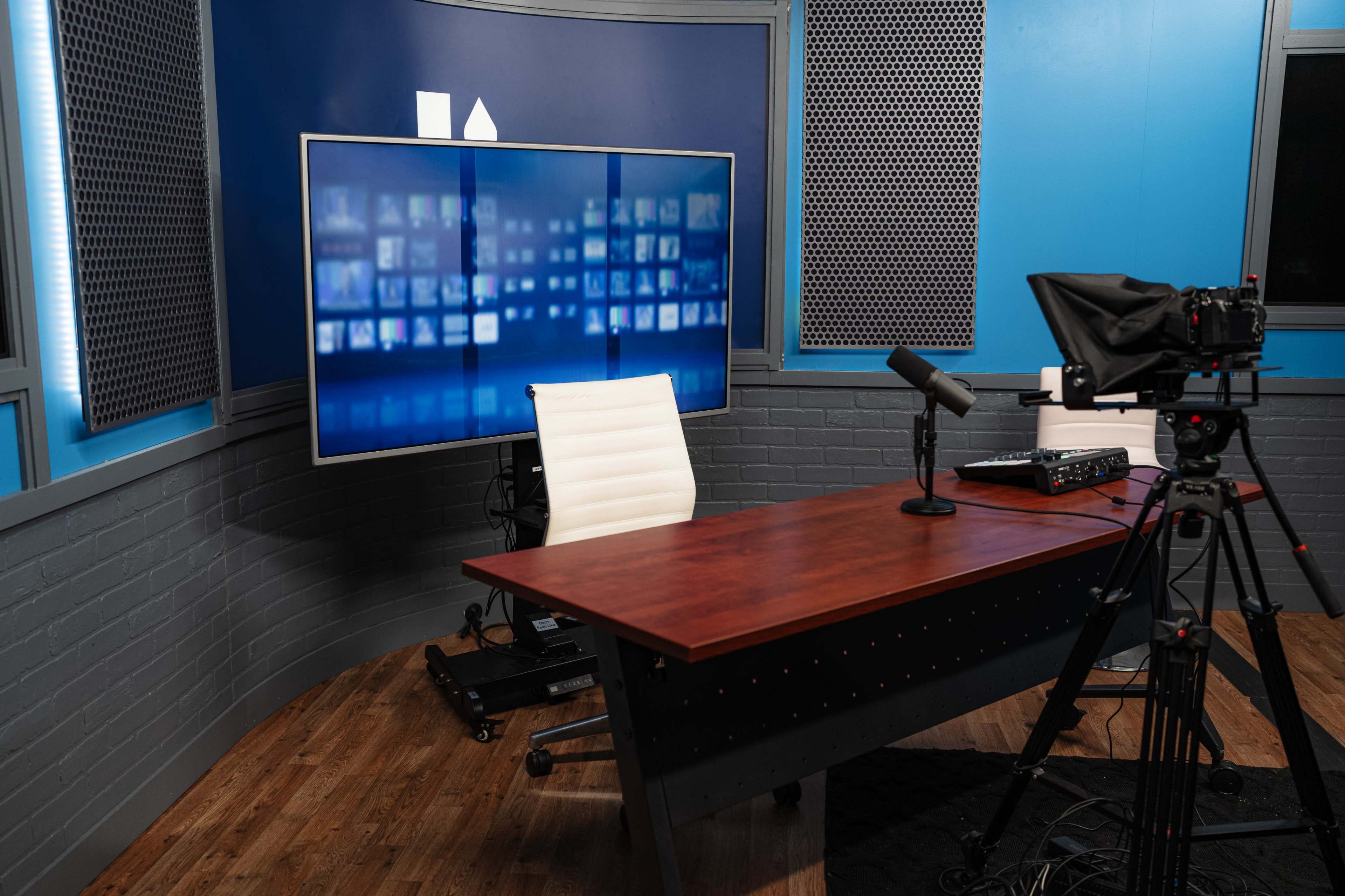 A modern studio setup featuring a wooden desk, a white swivel chair, a microphone, and a large screen displaying digital content.