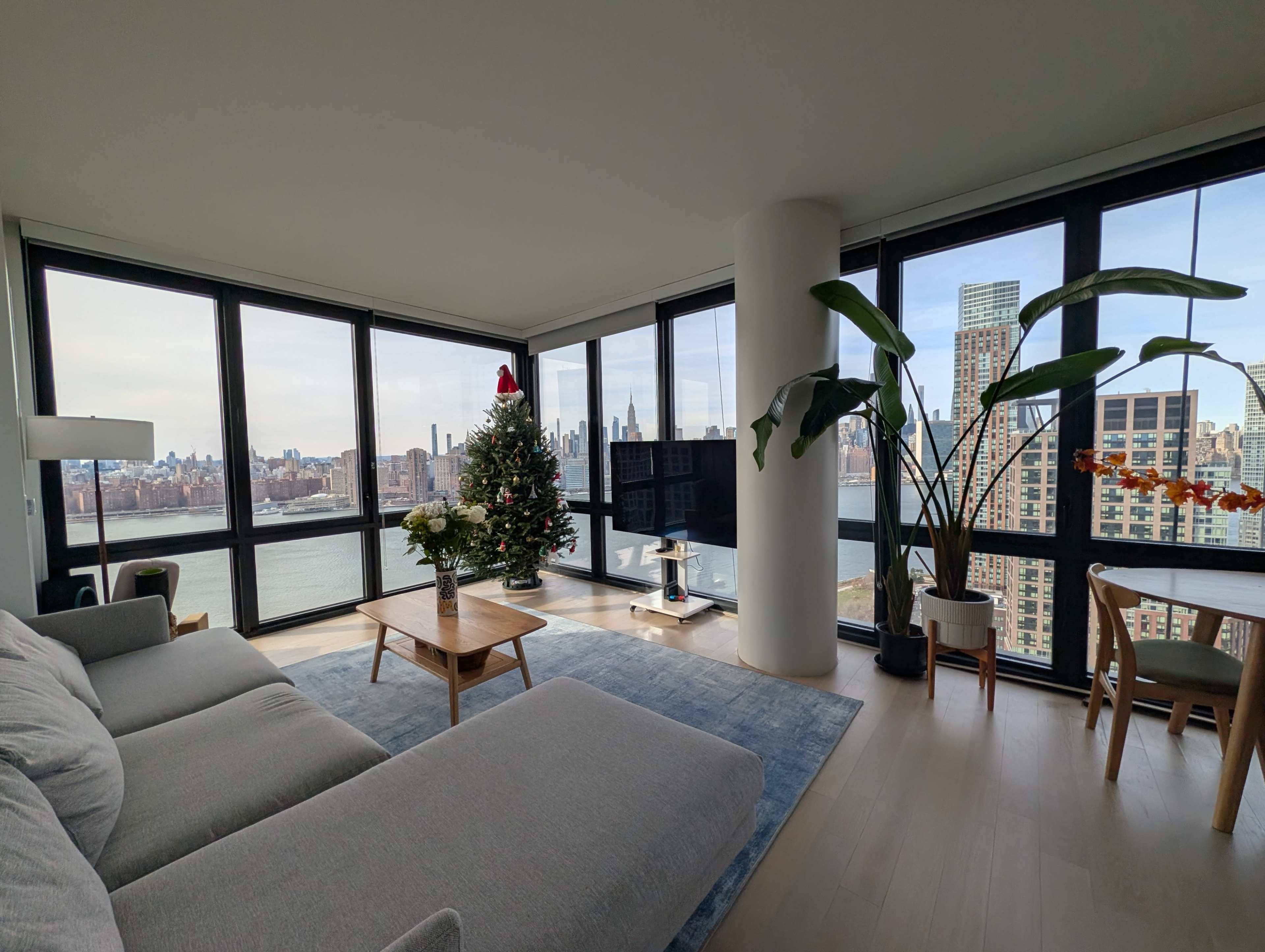 The image shows a modern living room with large windows offering a view of the city skyline and a Christmas tree in the corner.