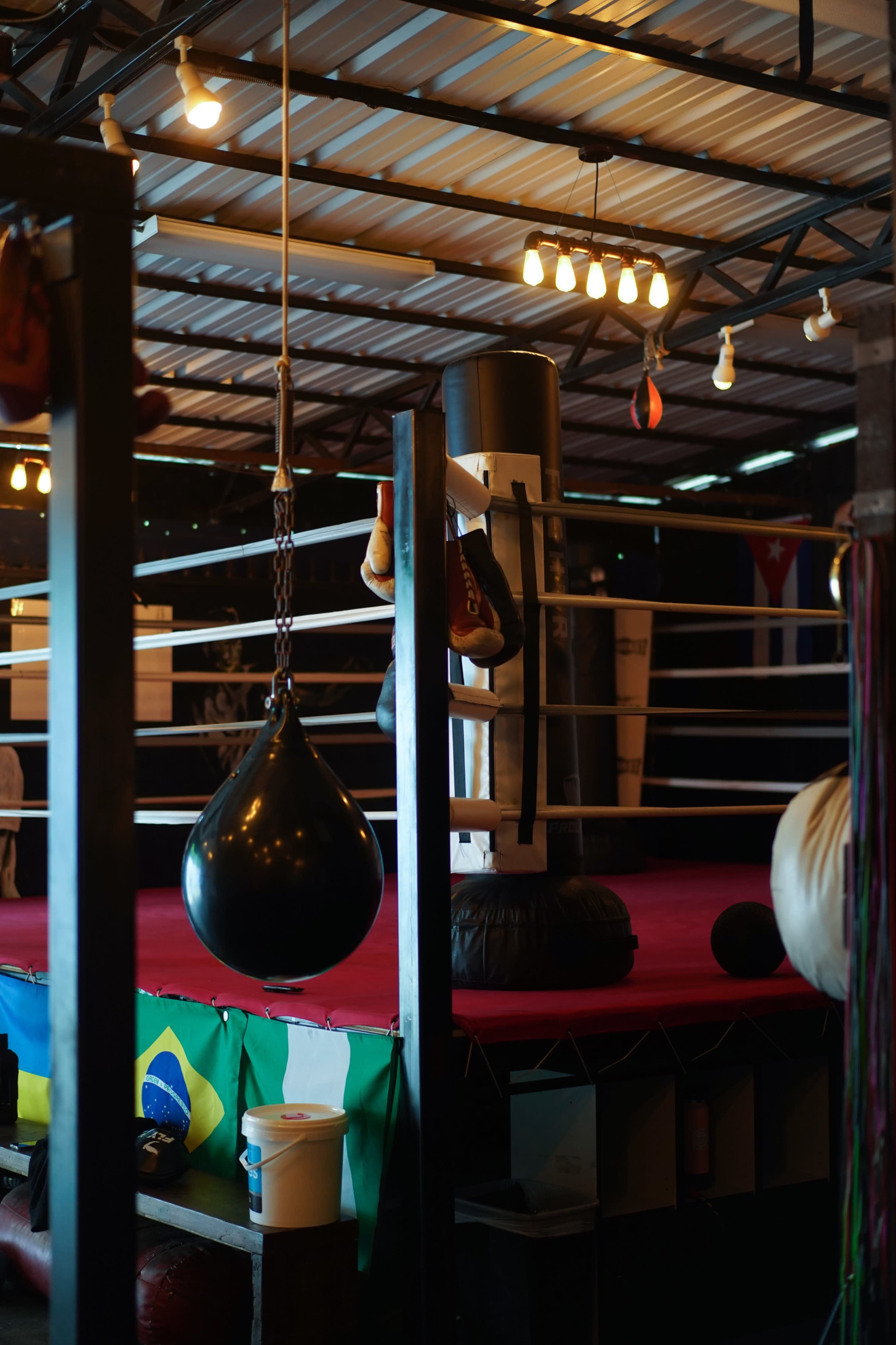 A boxing ring with red flooring and equipment in a gym setting, illuminated by hanging lights.