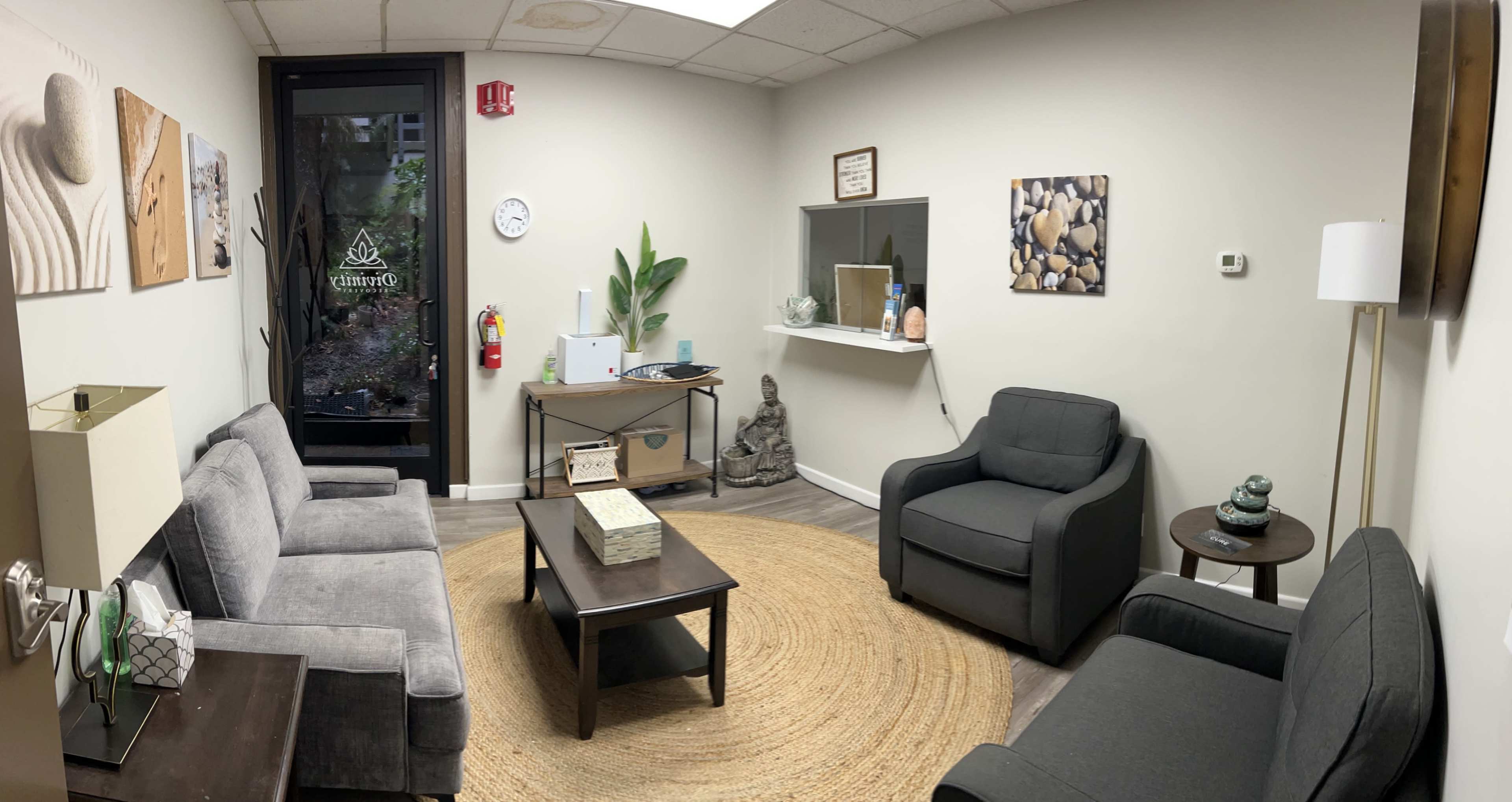 The image shows a waiting room with gray sofas, a round rug, a coffee table, and decorative items, including a small statue and plants, alongside a glass door leading outside.
