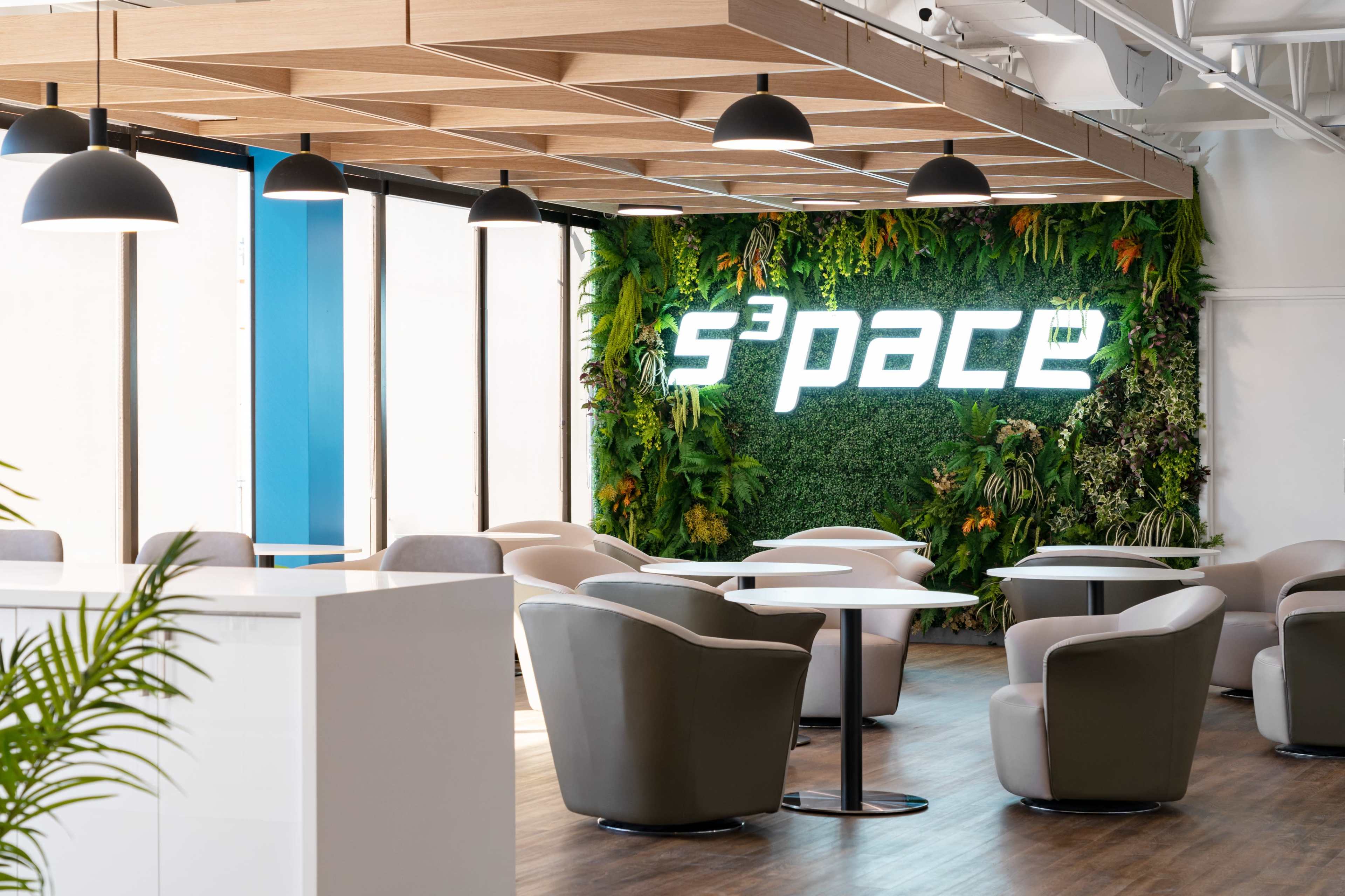 The image shows a modern indoor seating area featuring a green wall with the word "s³pace" illuminated, surrounded by comfortable chairs and tables.