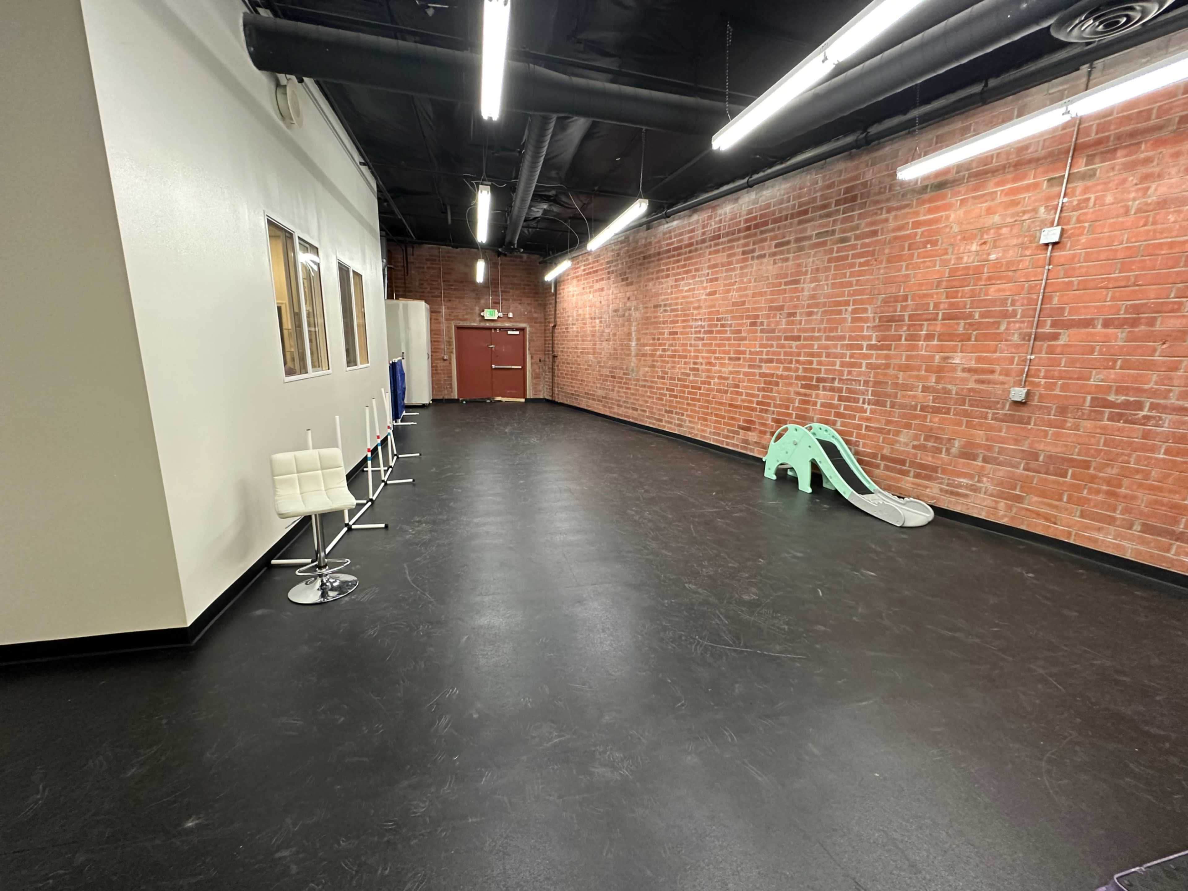 The room features a brick wall, a series of white bar stools, and a children's slide in a large, empty space with black flooring and overhead fluorescent lights.