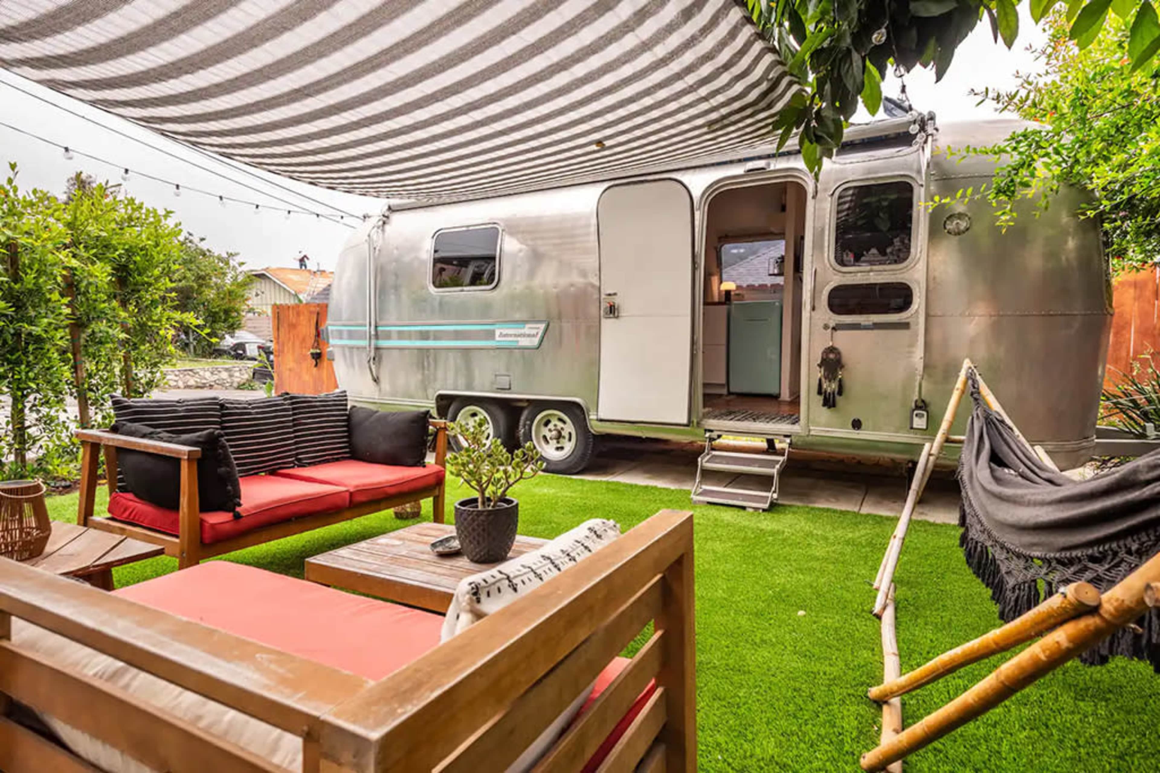 A silver Airstream trailer is parked next to a cozy outdoor seating area with red cushions on a faux grass surface, surrounded by greenery and a striped canopy overhead.