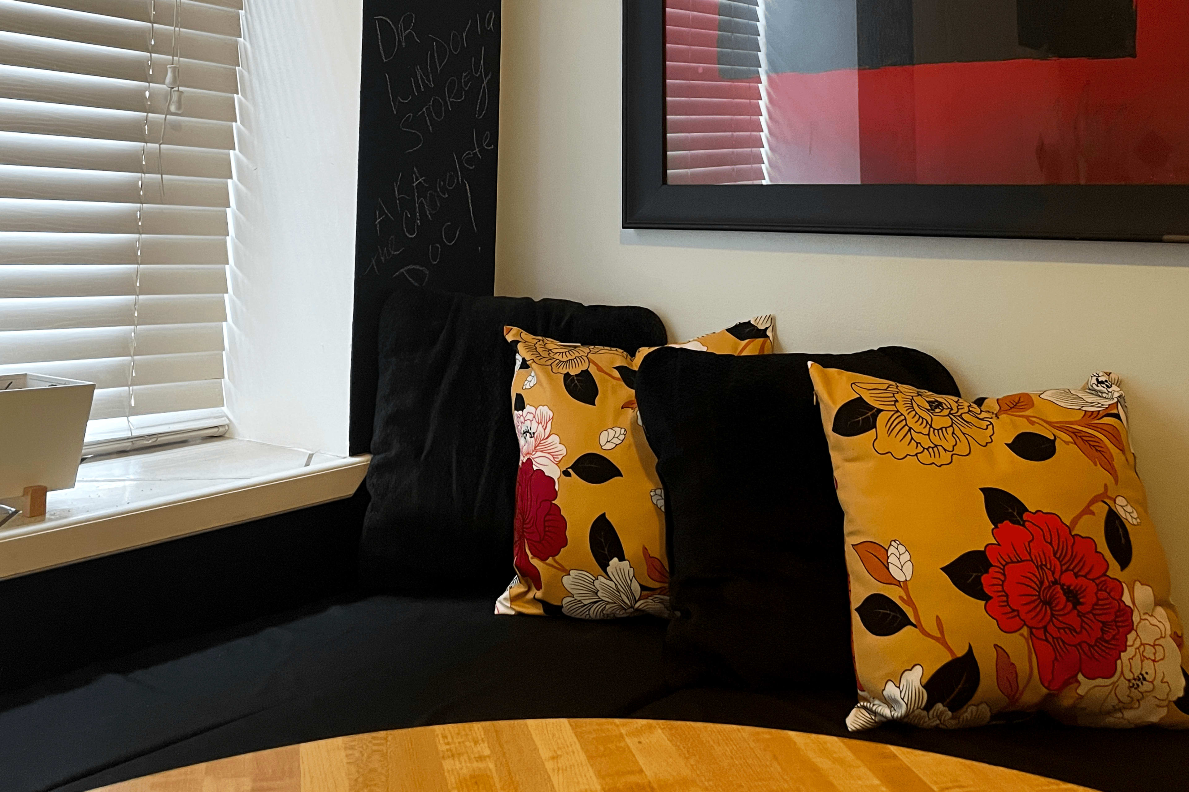 A corner seating area features a wooden table and decorative pillows with floral patterns against a backdrop of a black and red framed artwork.