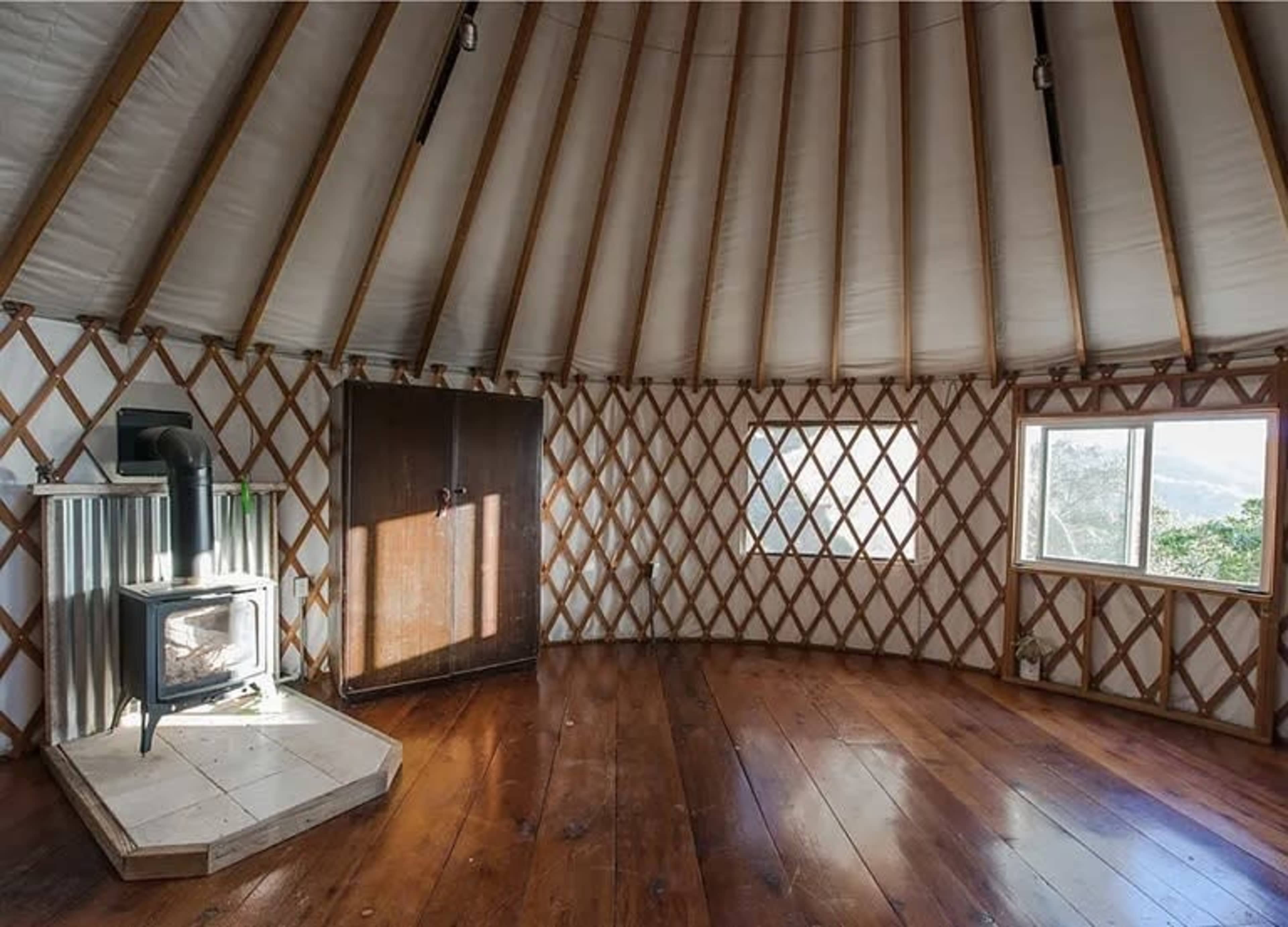 The interior of a yurt features wooden walls with a circular design, a wood-burning stove, and large windows providing natural light.