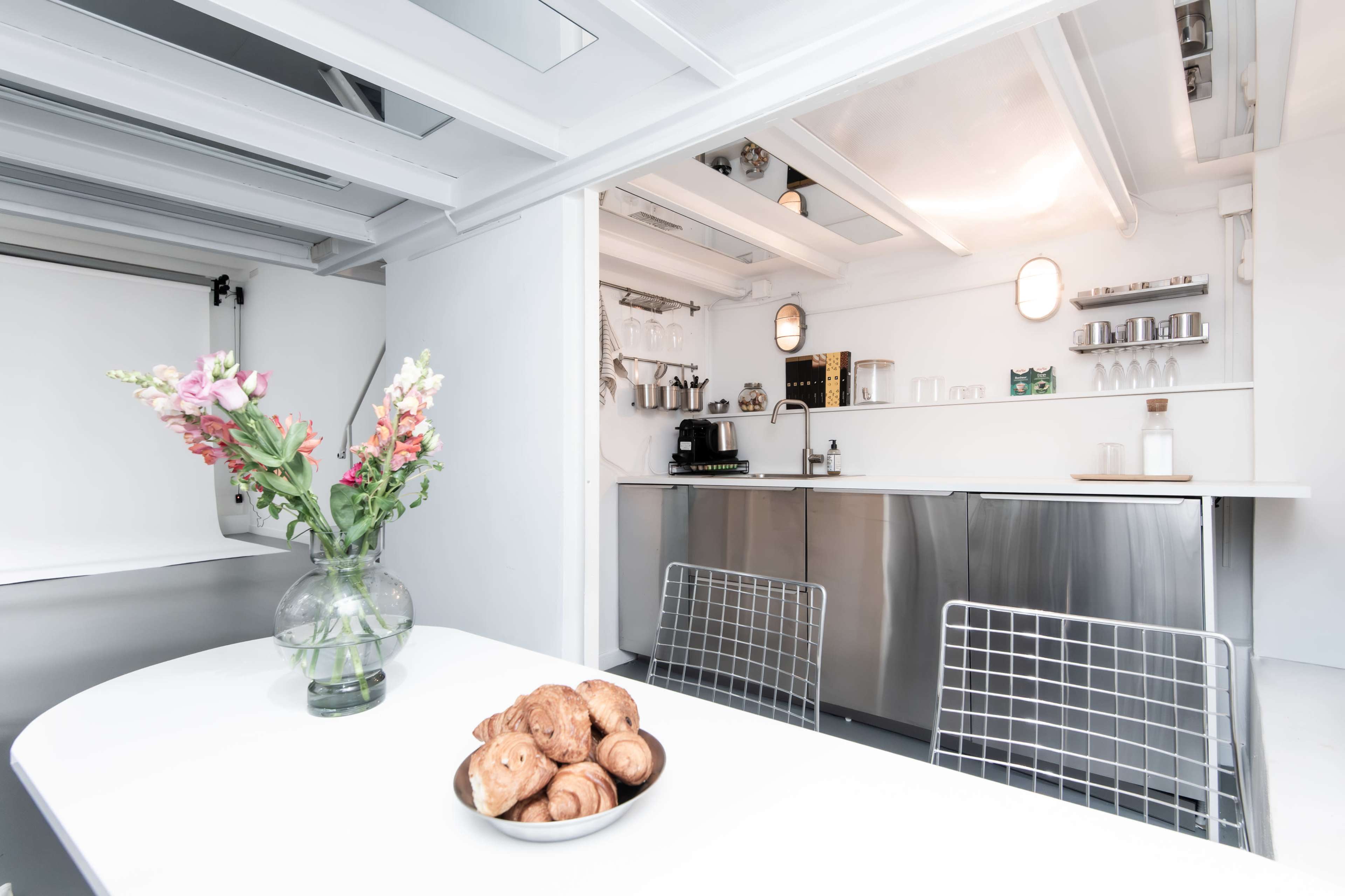 A modern kitchen features a table with a bowl of pastries and a vase of flowers, with stainless steel appliances and minimal decor in the background.