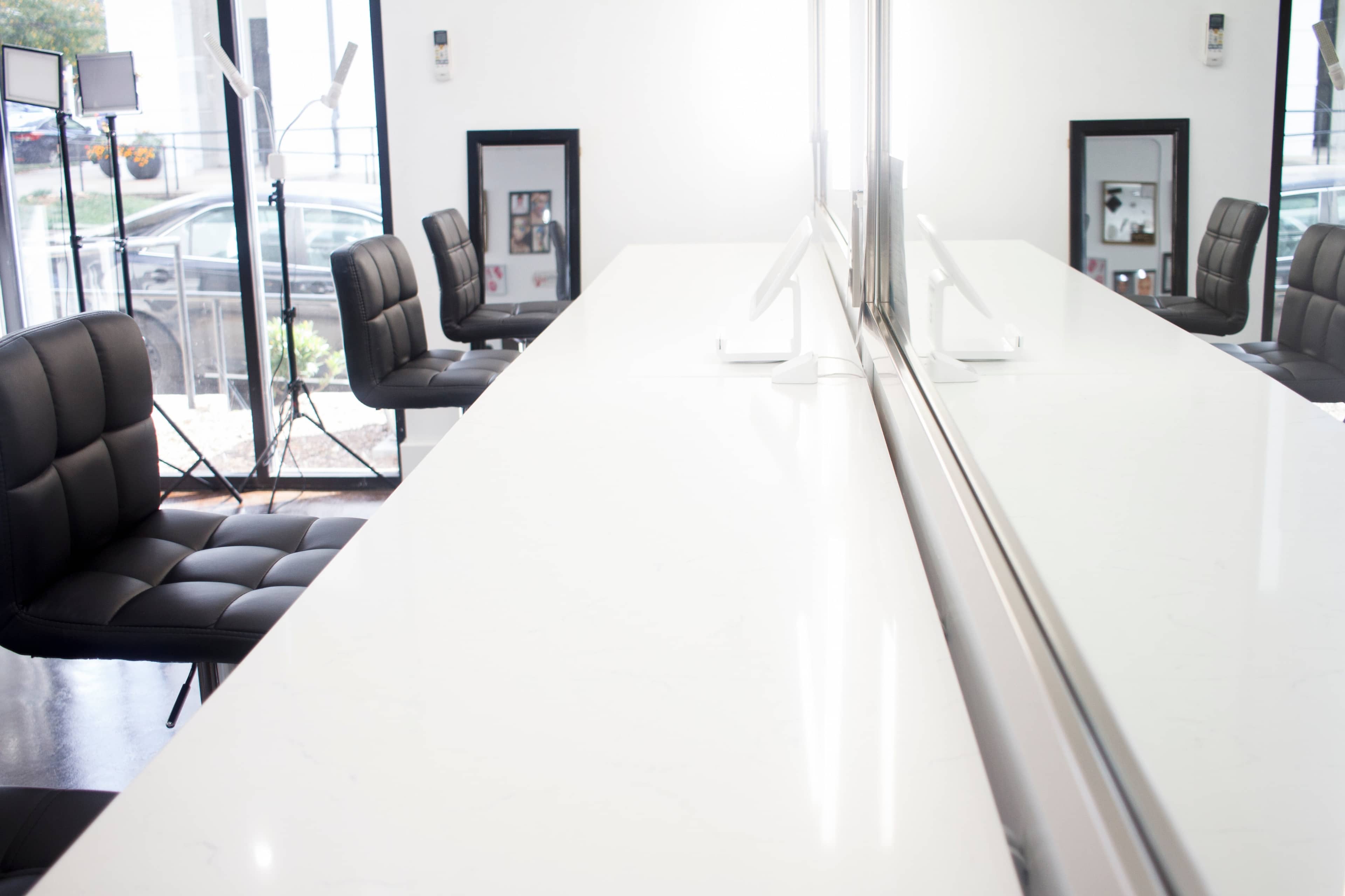 A modern salon interior featuring a long white countertop with two black salon chairs positioned on either side, reflecting a mirror across the surface.