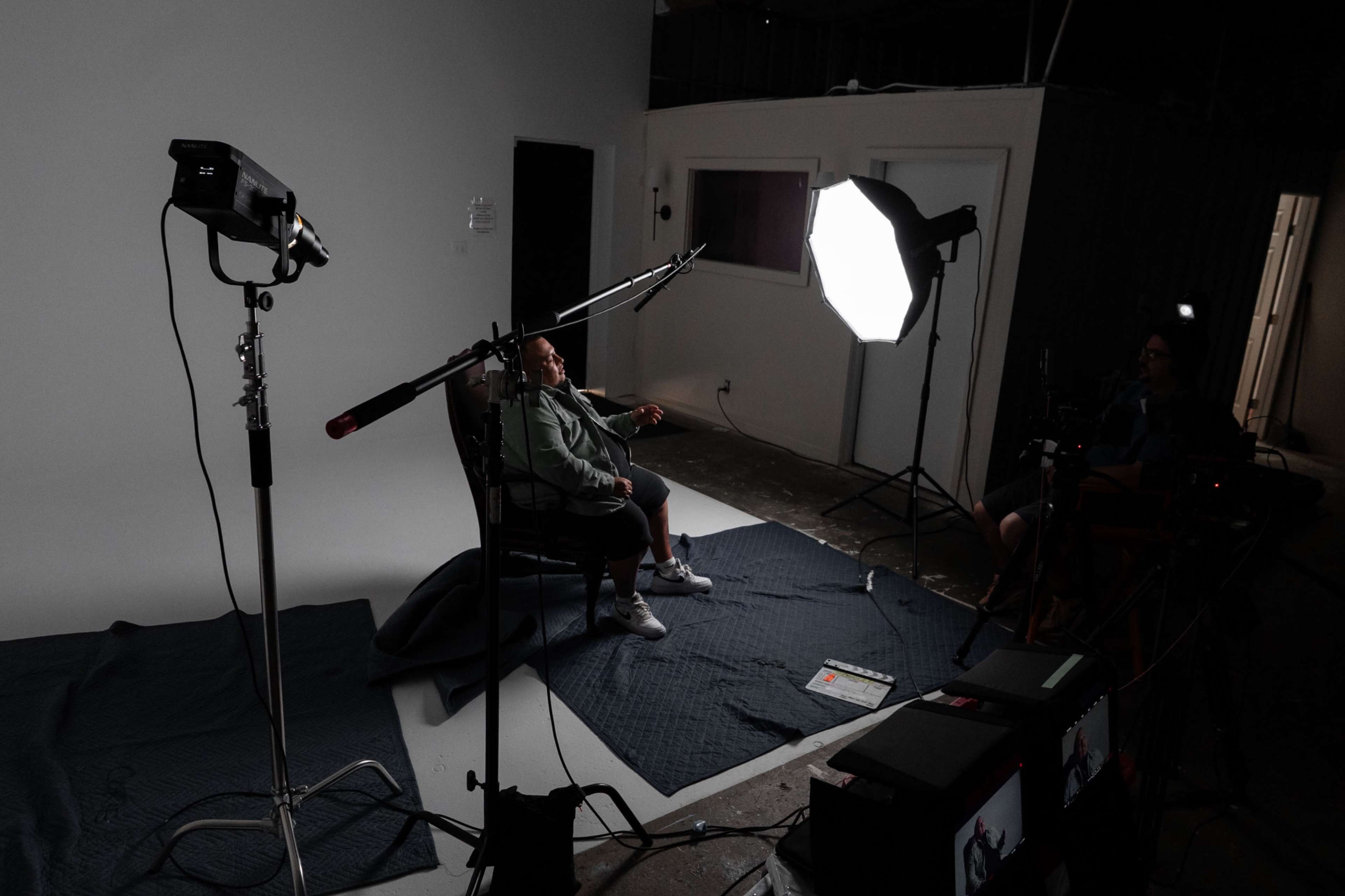 A person sits in a chair in a photography studio, surrounded by lighting equipment and a plain backdrop.