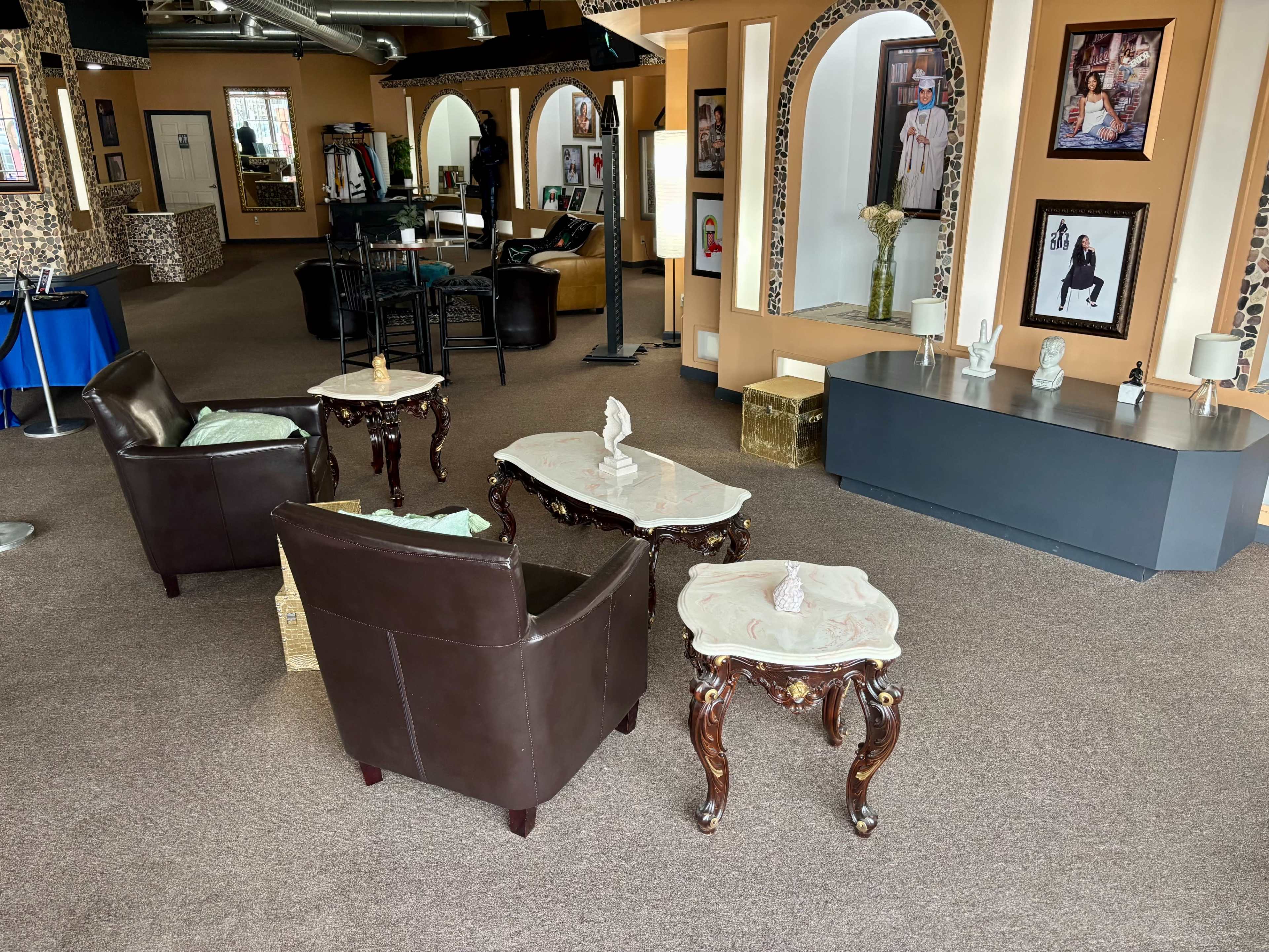 The scene features a spacious room with two brown leather chairs, two ornate tables with marble tops, and a grey sideboard, surrounded by framed artwork and a decorative wall.