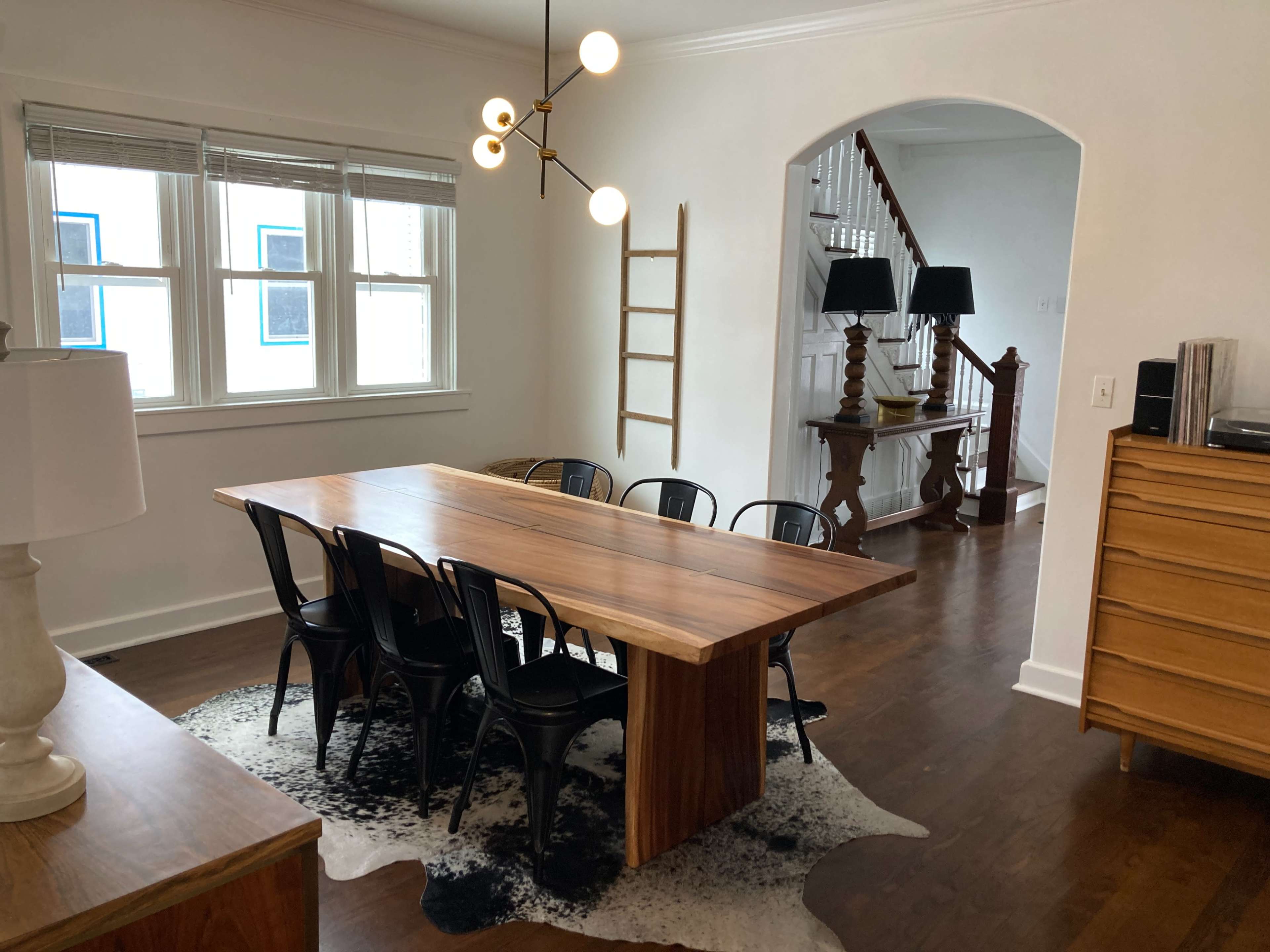 A modern dining area featuring a long wooden table surrounded by black chairs, with large windows letting in natural light and a staircase visible in the background.