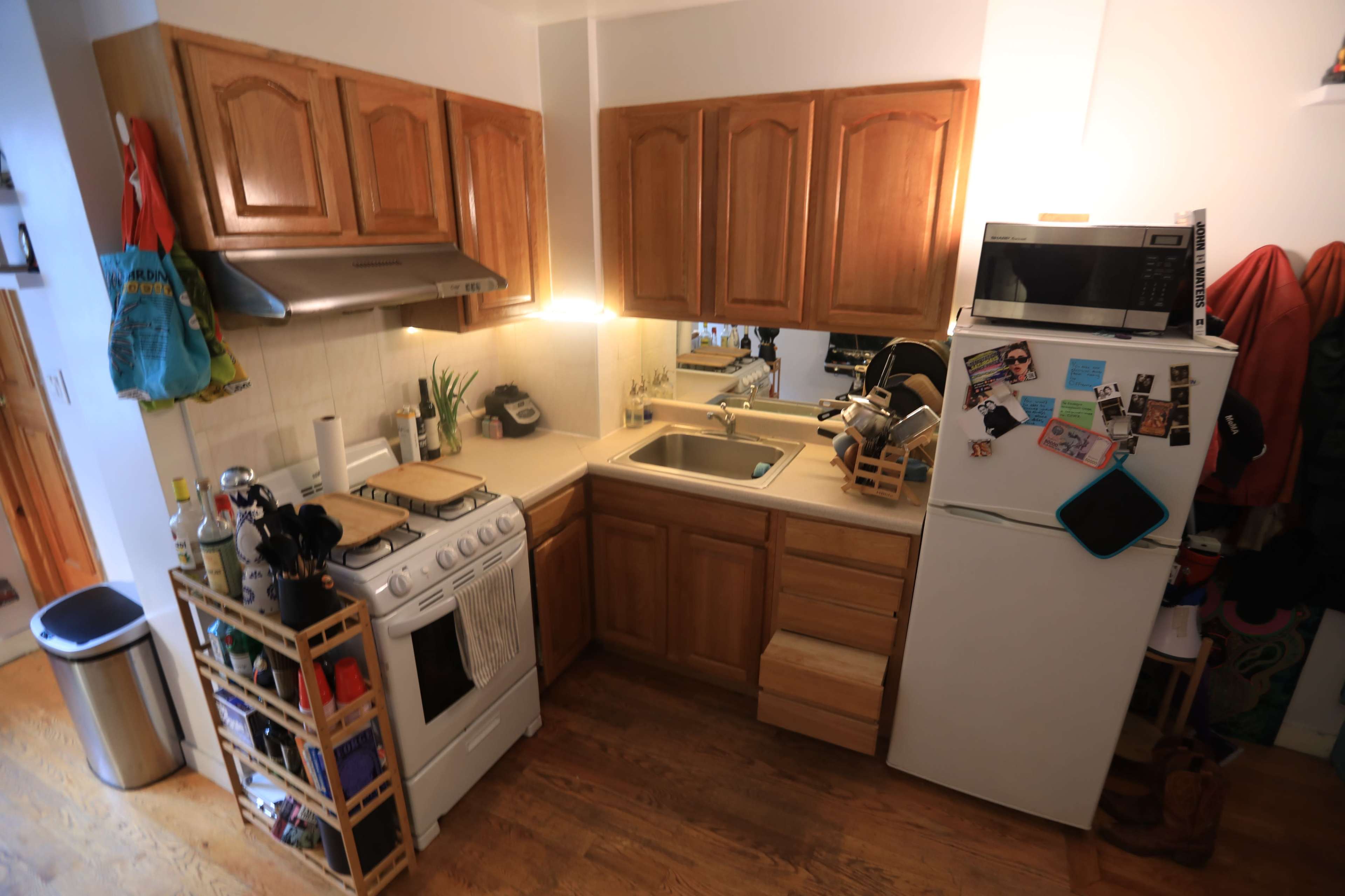 A compact kitchen features wooden cabinets, a stove, a sink, a refrigerator, and various kitchen items arranged on shelves.