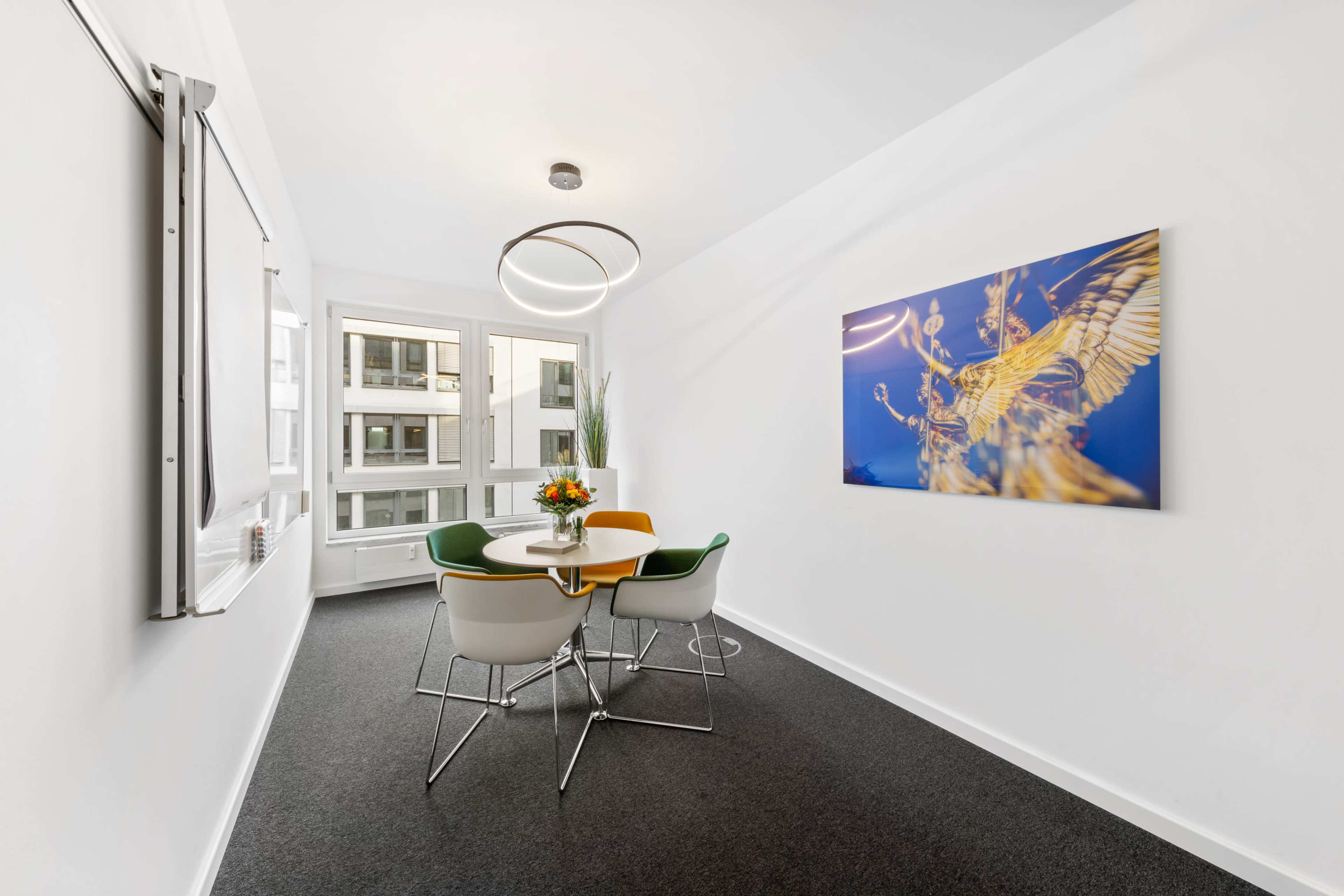 A well-lit meeting room features a round table with colorful chairs, a floral centerpiece, a wall-mounted screen, and a framed artwork.