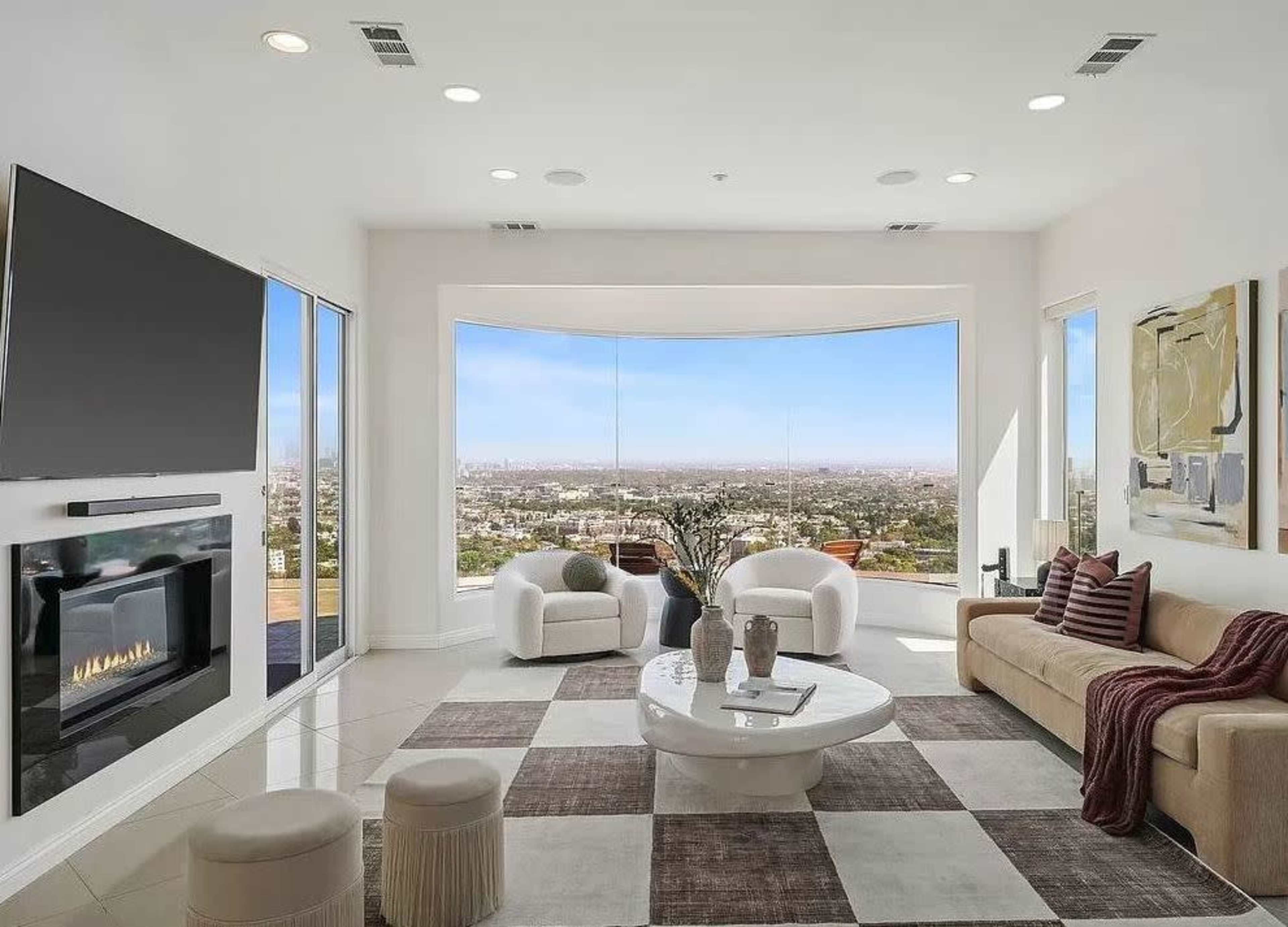The image shows a modern living room with large windows offering a city view, a beige sofa, two white armchairs, and a round coffee table on a patterned rug.