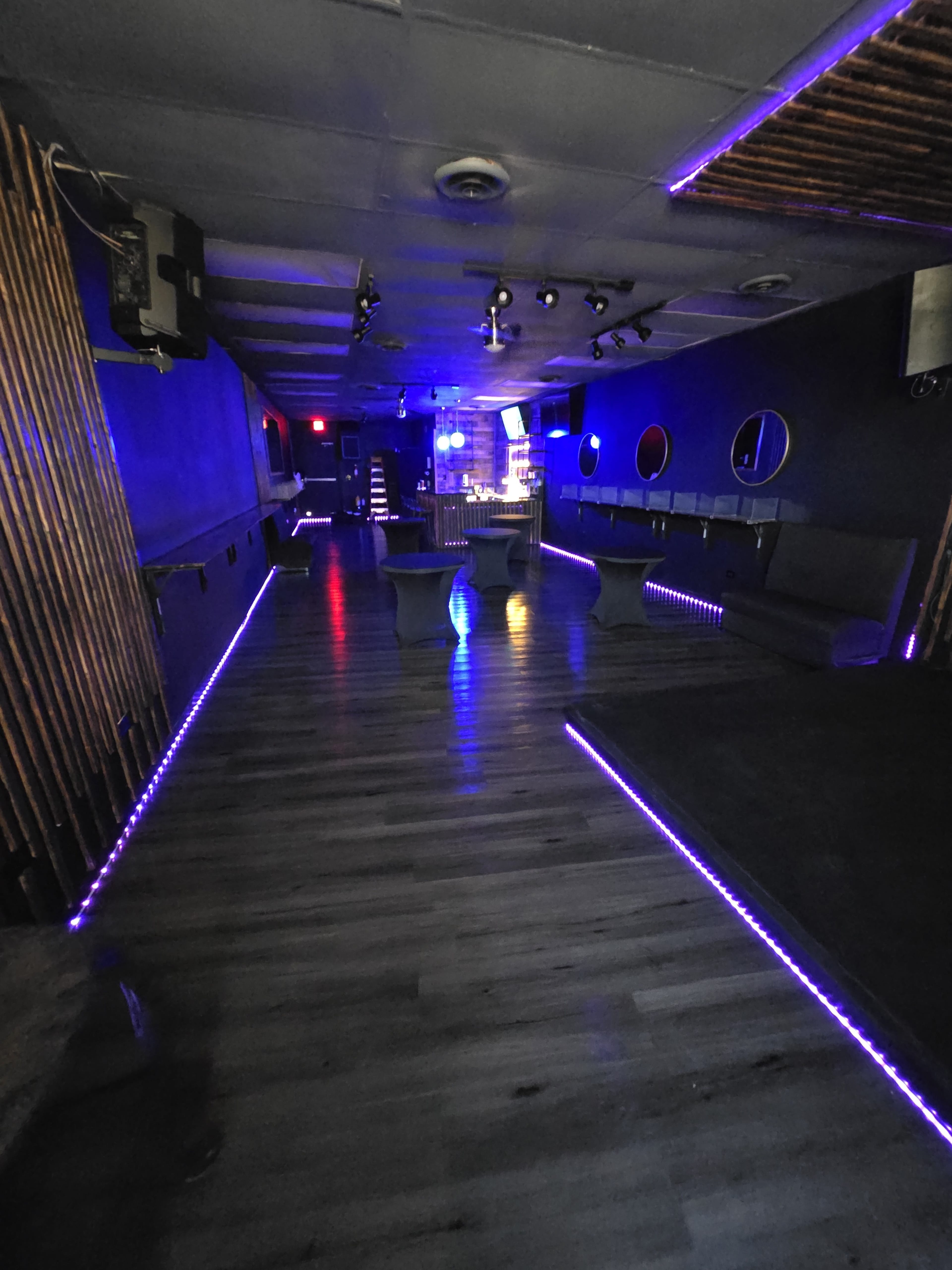 The interior of a dimly lit nightclub features sleek seating areas, illuminated by purple LED lights along the perimeter.