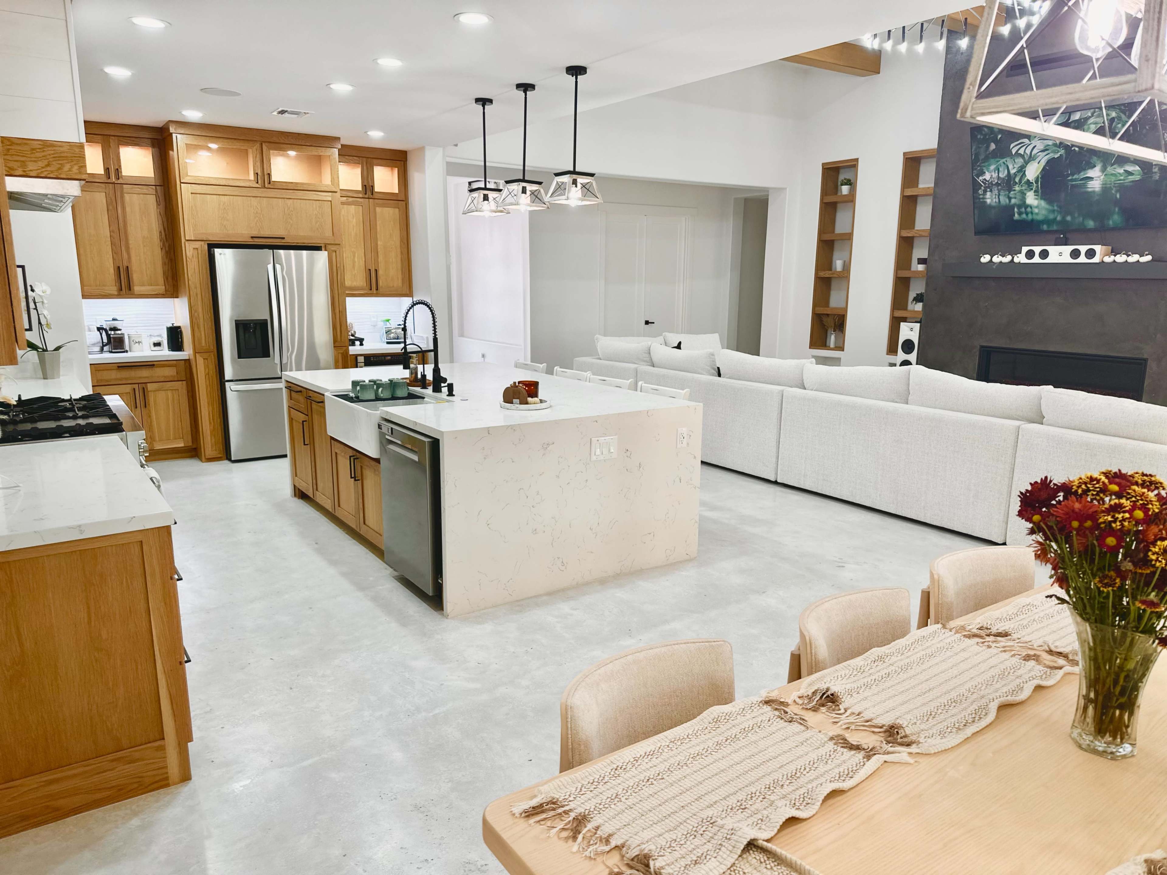 The image shows a modern kitchen and living area with wooden cabinetry, a large island, and a sectional sofa arranged around a fireplace.