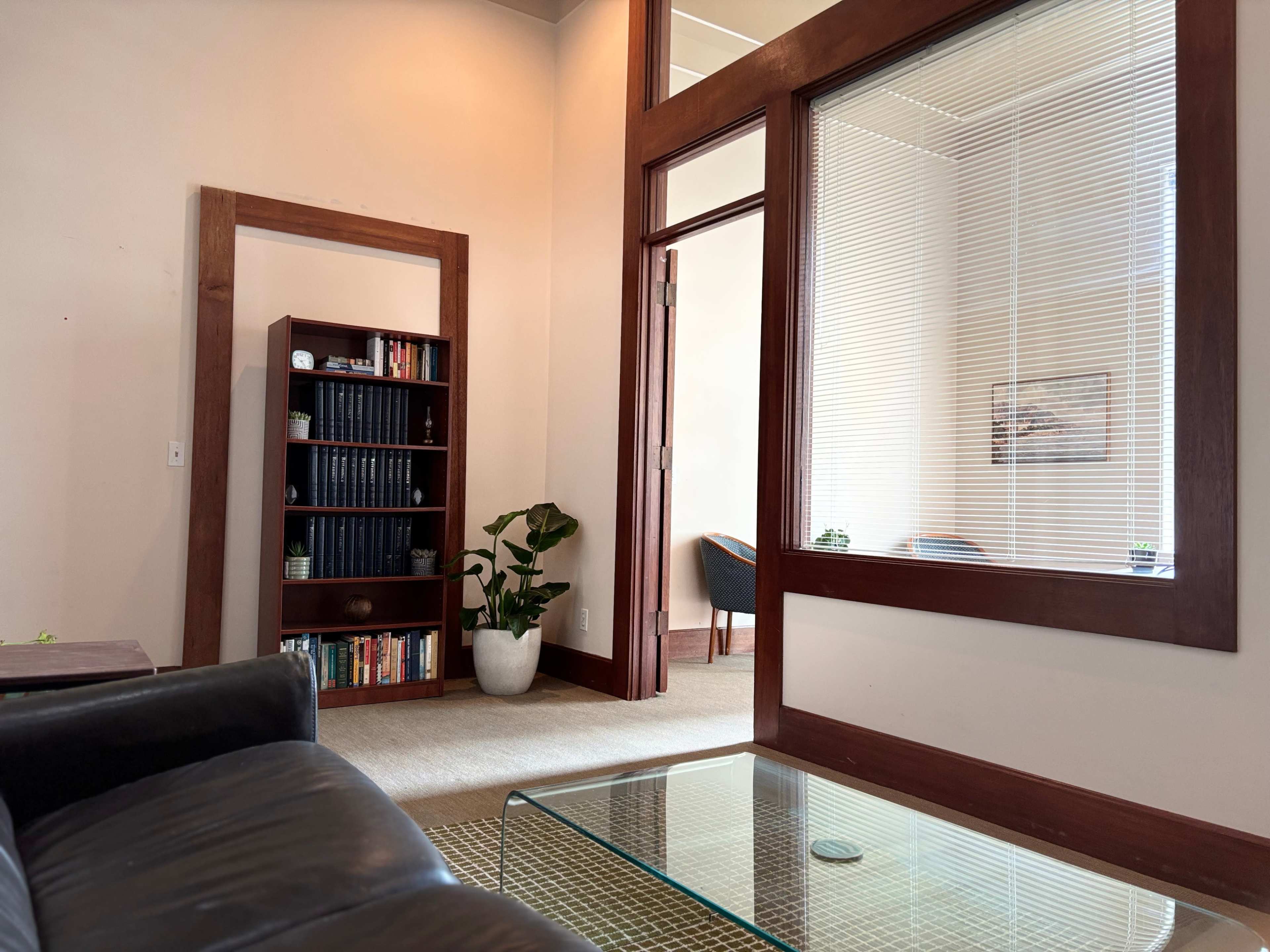 The image shows a well-lit office space with a black couch, a glass coffee table, a bookshelf filled with books, and a large window between two rooms.