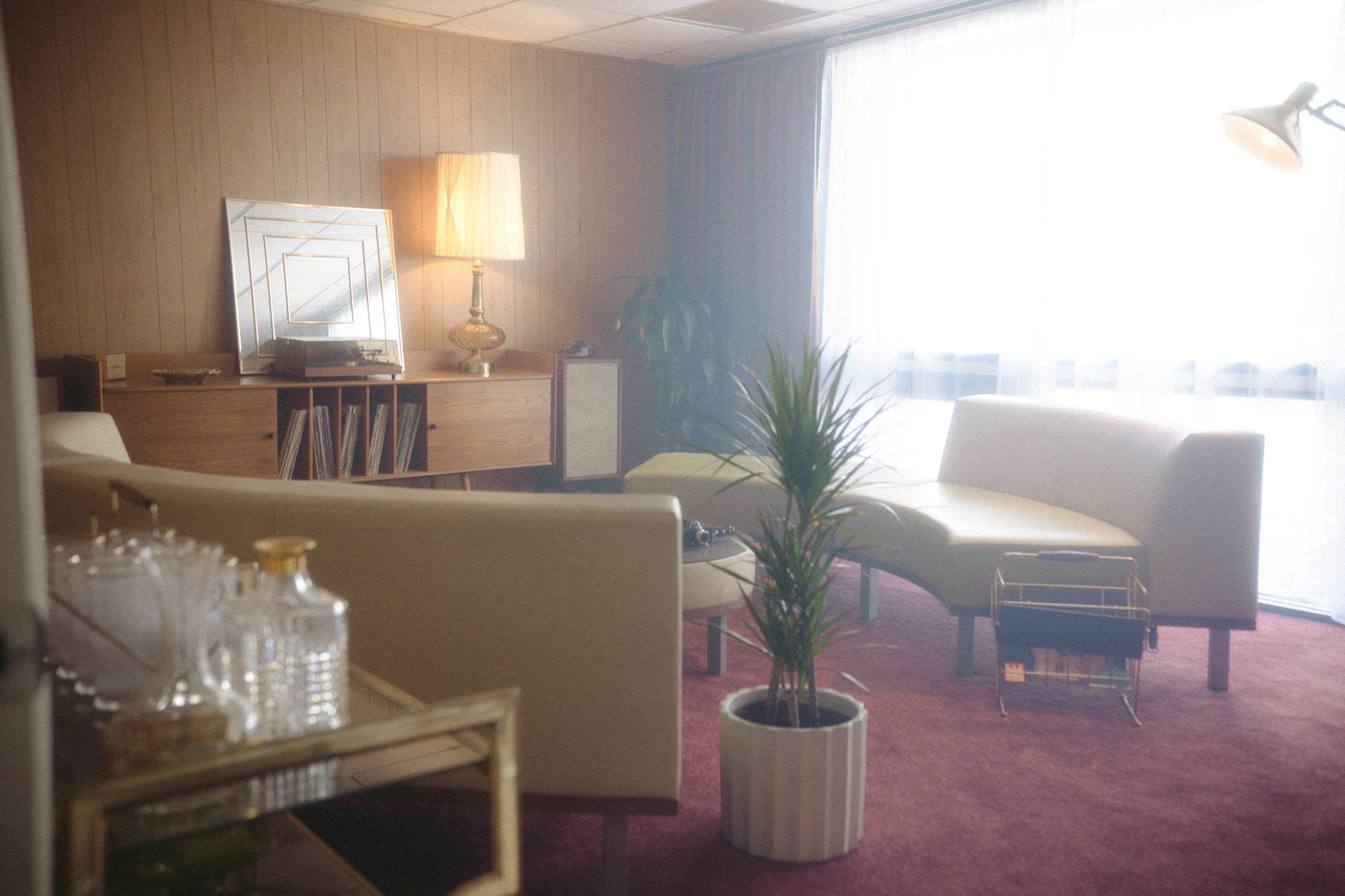 The image shows a vintage-style living room with two curved sofas, a tall lamp, a potted plant, and a wooden cabinet, all under soft lighting.