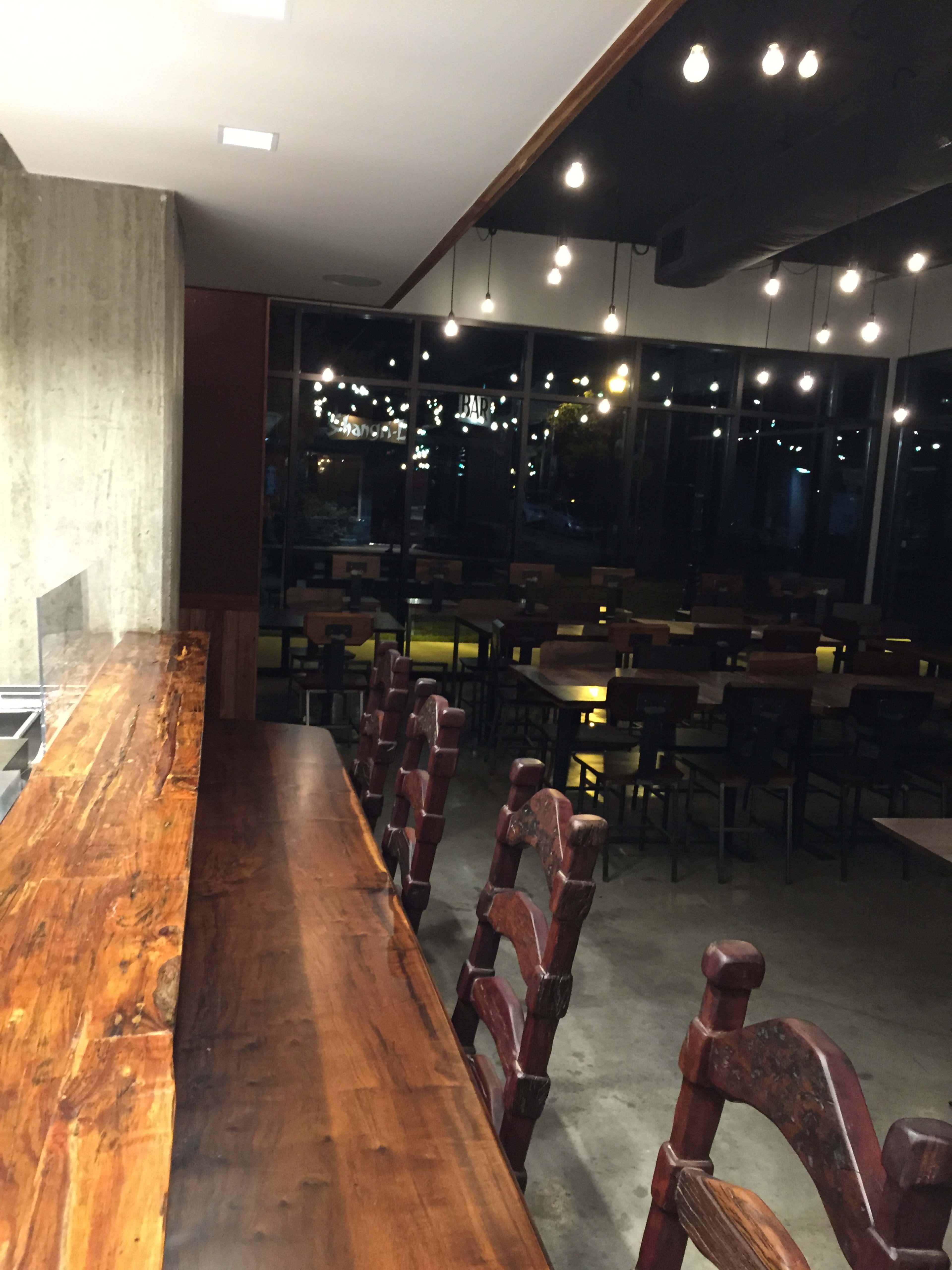 The image shows a modern, empty restaurant interior with wooden tables and a long wooden counter illuminated by overhead lights.