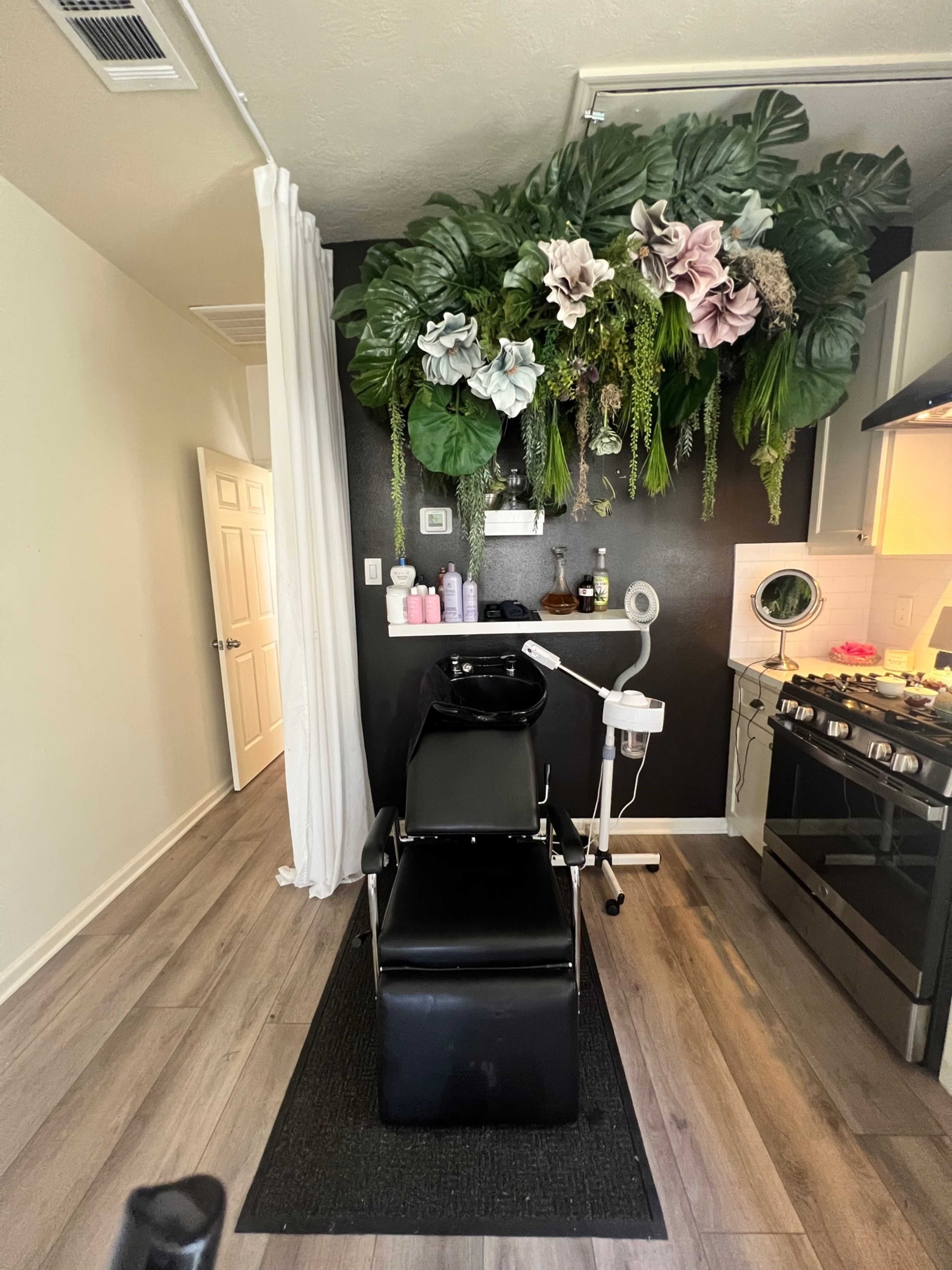 The image shows a hair salon area with a black shampoo chair, a sink, and a wall adorned with large artificial plants.