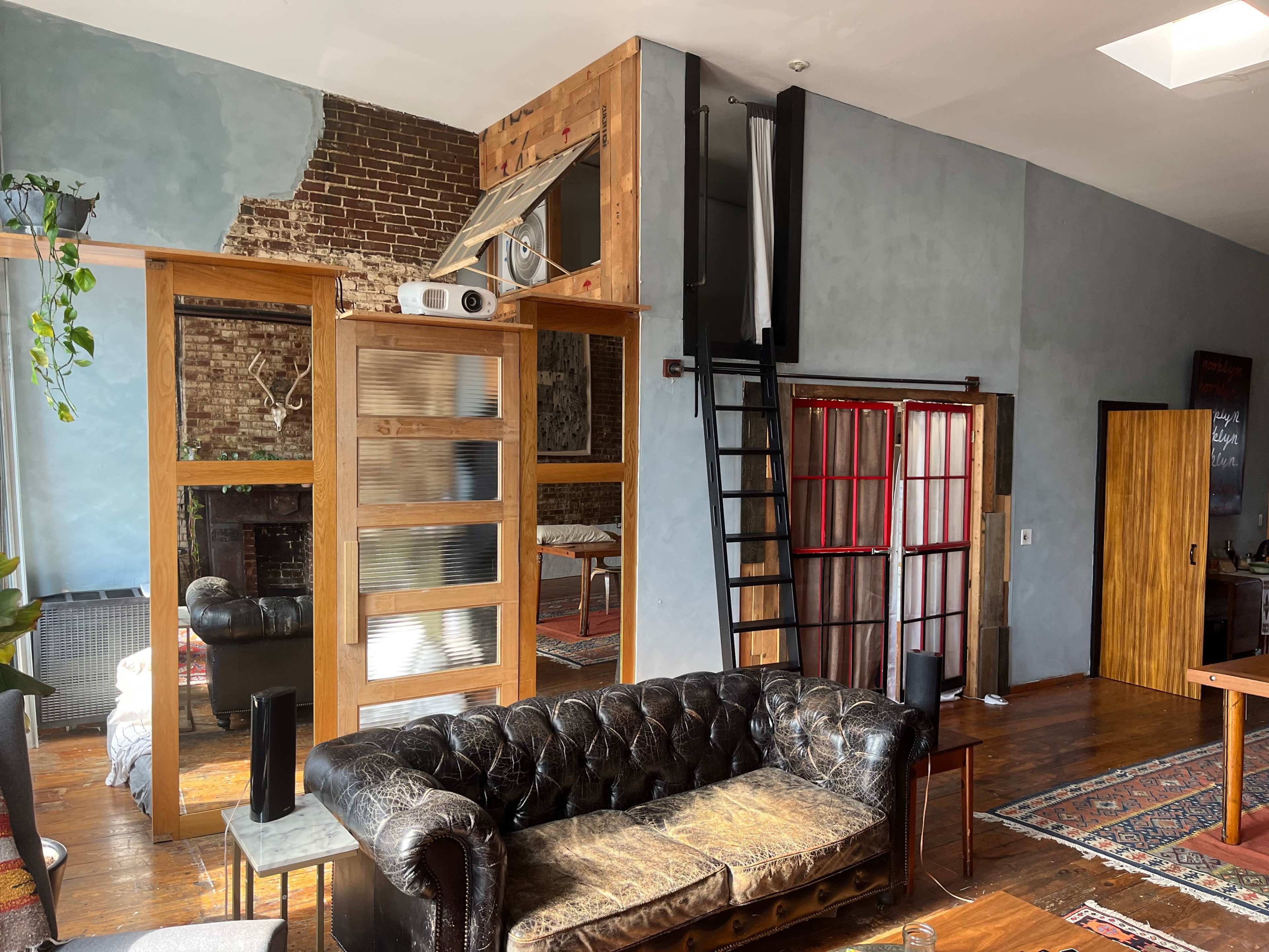 A stylish loft living area with a black leather chesterfield sofa, exposed brick walls, wooden accents, and a metal staircase leading to a higher level.