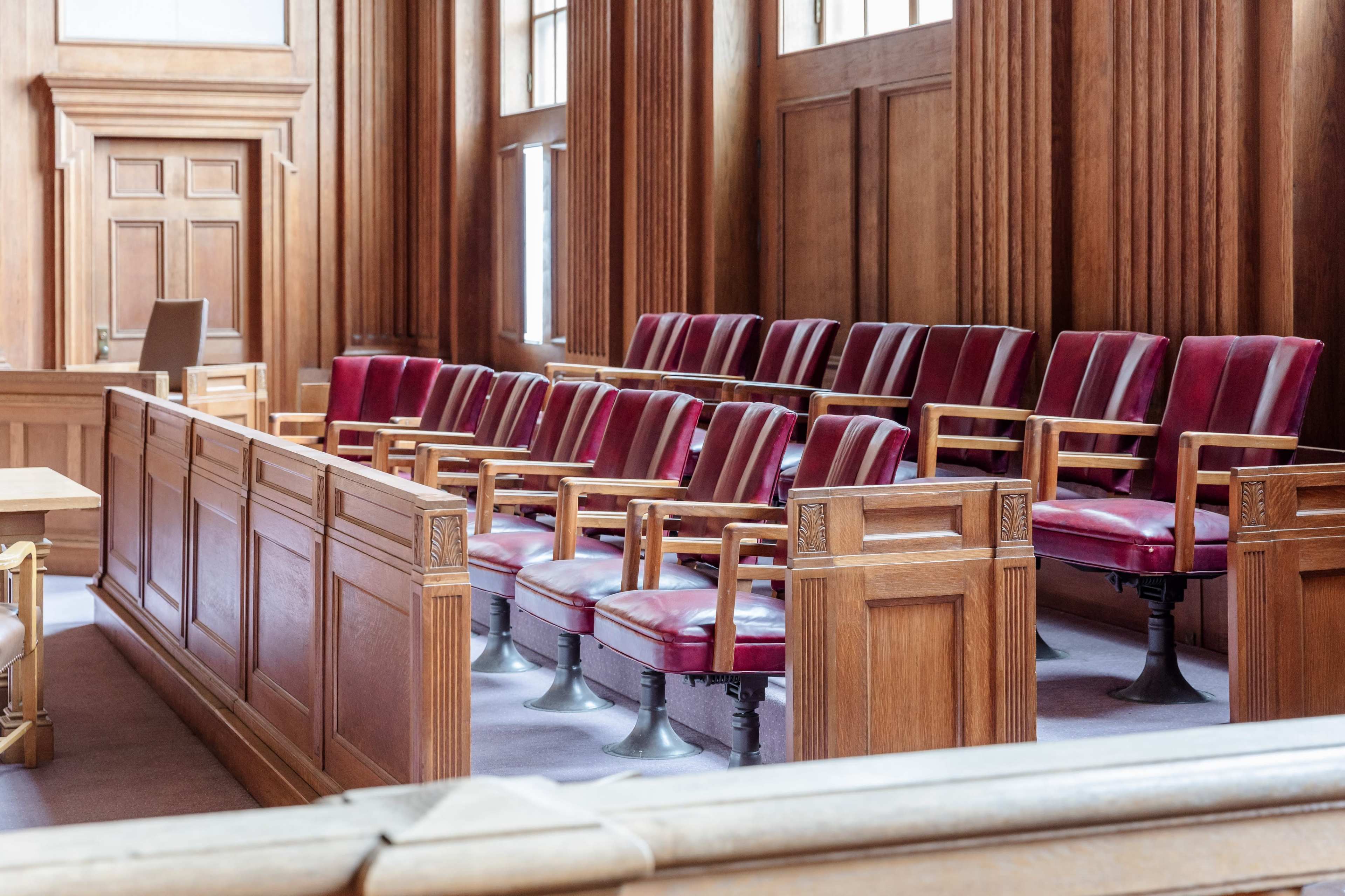 The image shows empty wooden jury seats arranged in a courtroom with large windows and a wooden panelled interior.