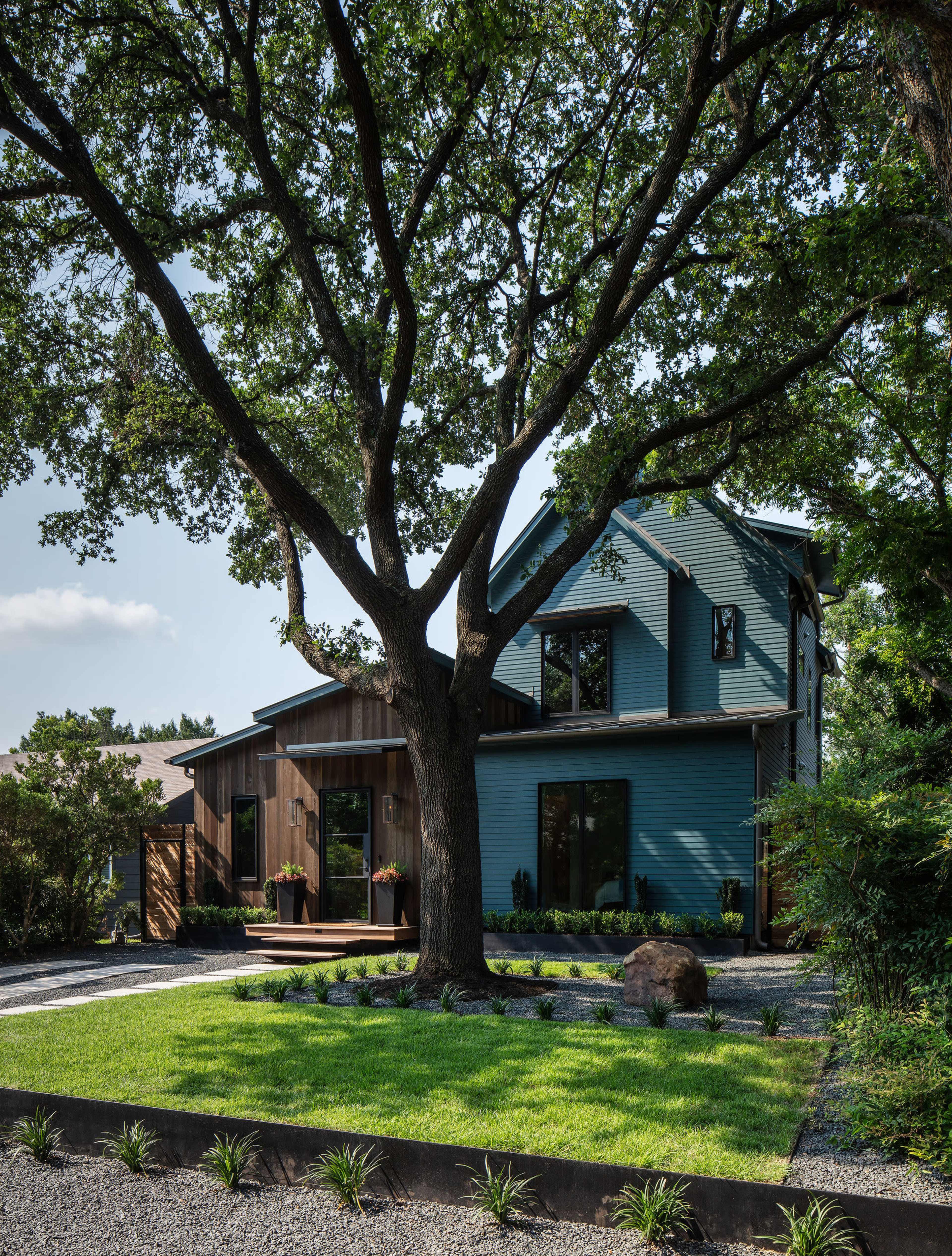 A modern two-story house with a combination of blue and wooden siding is set in a landscaped yard featuring a large tree, gravel pathways, and greenery.