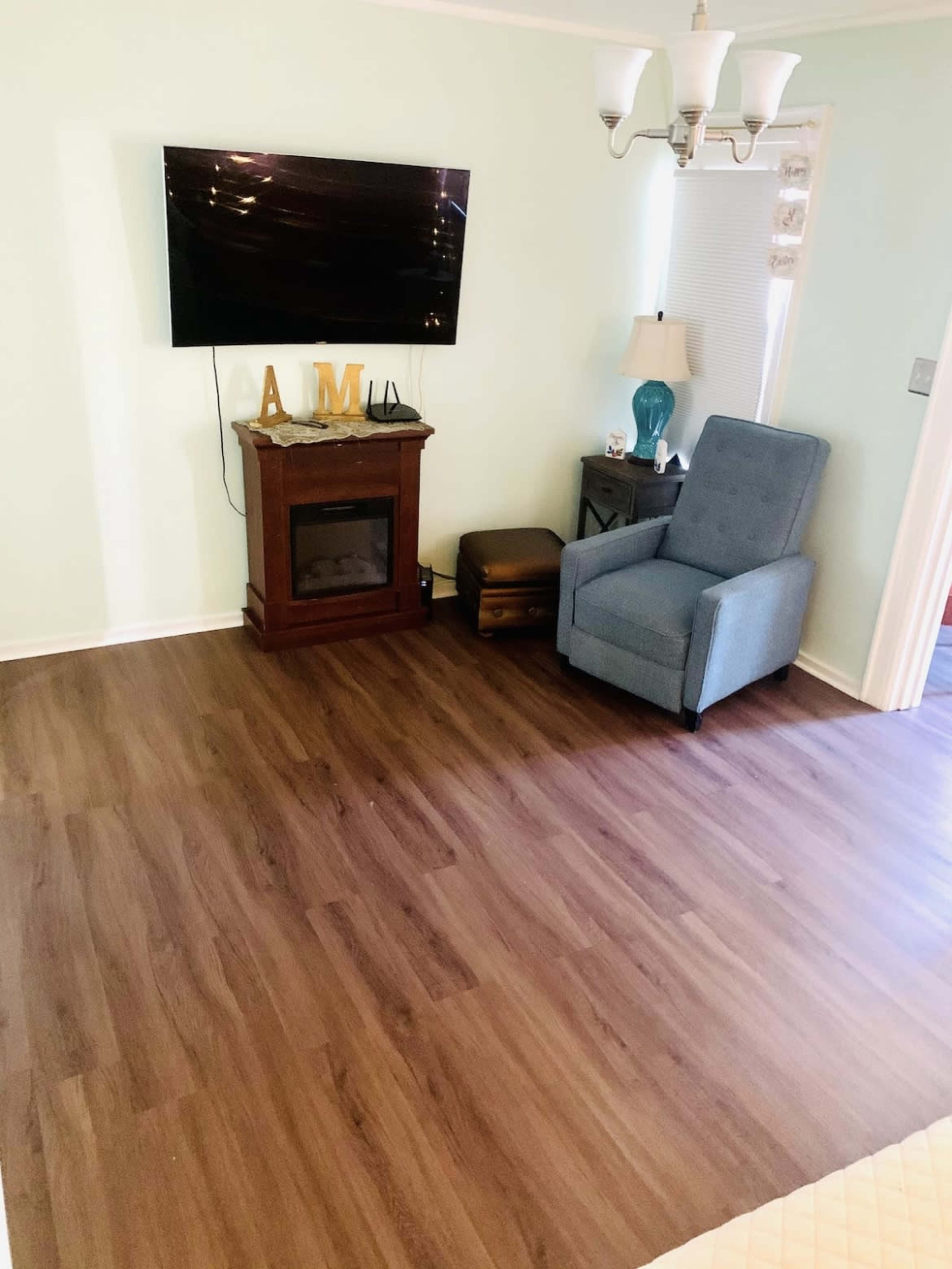 A living room with light green walls, a mounted television, a small electric fireplace, a blue armchair, and a light brown decorative box on the floor.