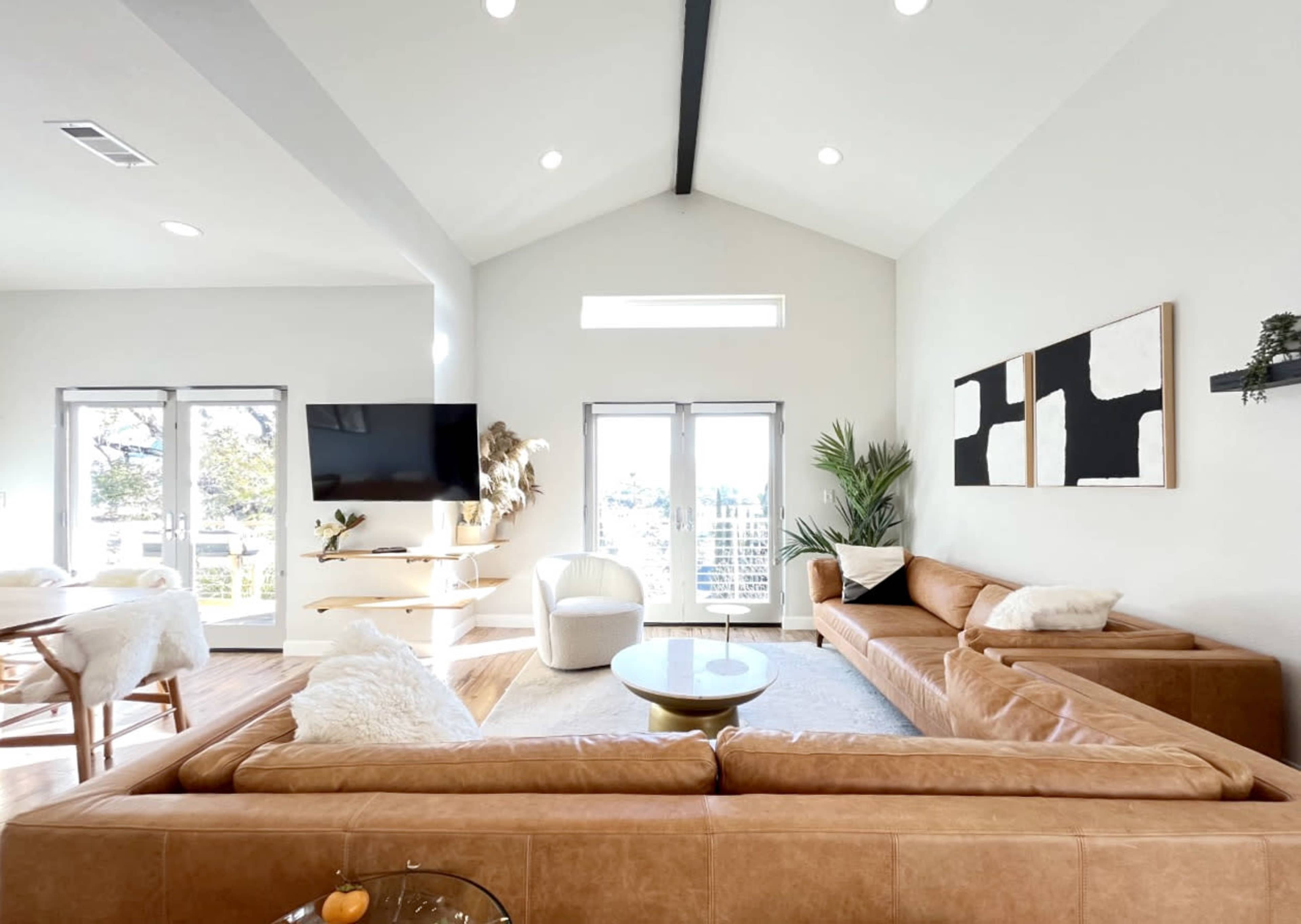A modern living room featuring a large, brown sectional sofa, a round coffee table, a wall-mounted TV, and large windows allowing natural light to enter.