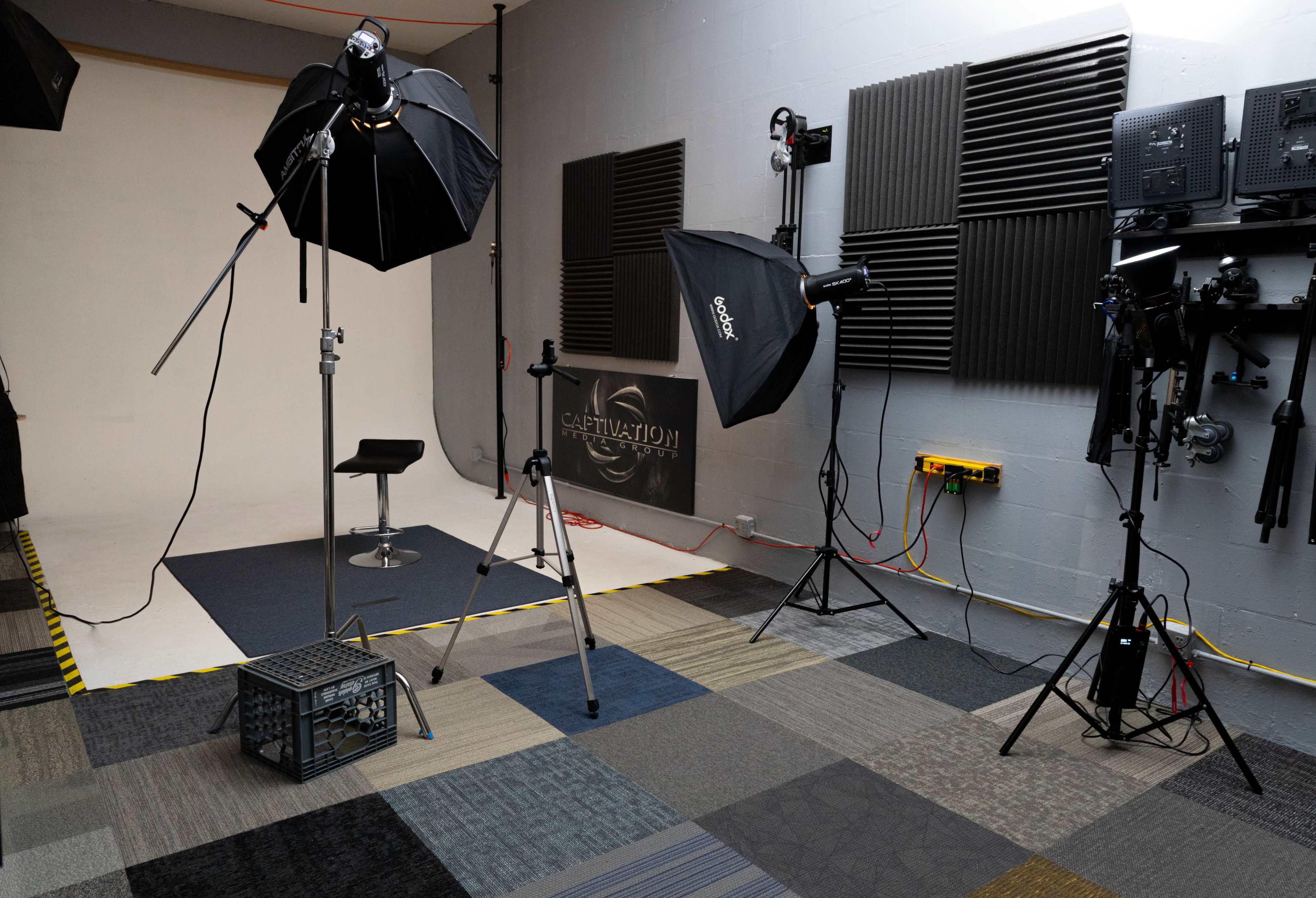 The image shows a photography studio setup with multiple lights, a stool, and soundproofing panels on the walls.