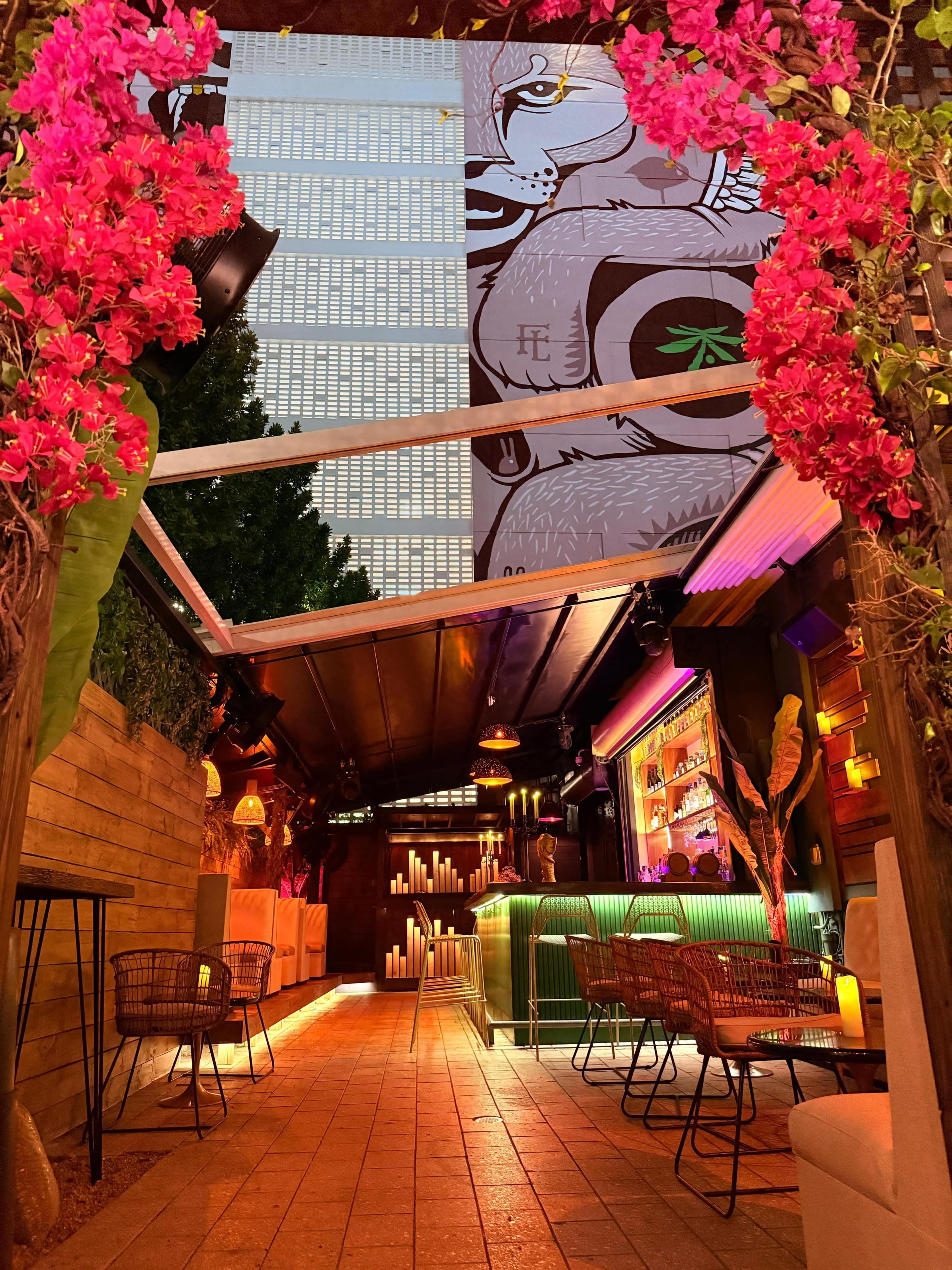The image depicts an outdoor bar area adorned with vibrant pink flowers, featuring a lit-up bar and a large mural on the wall in the background.