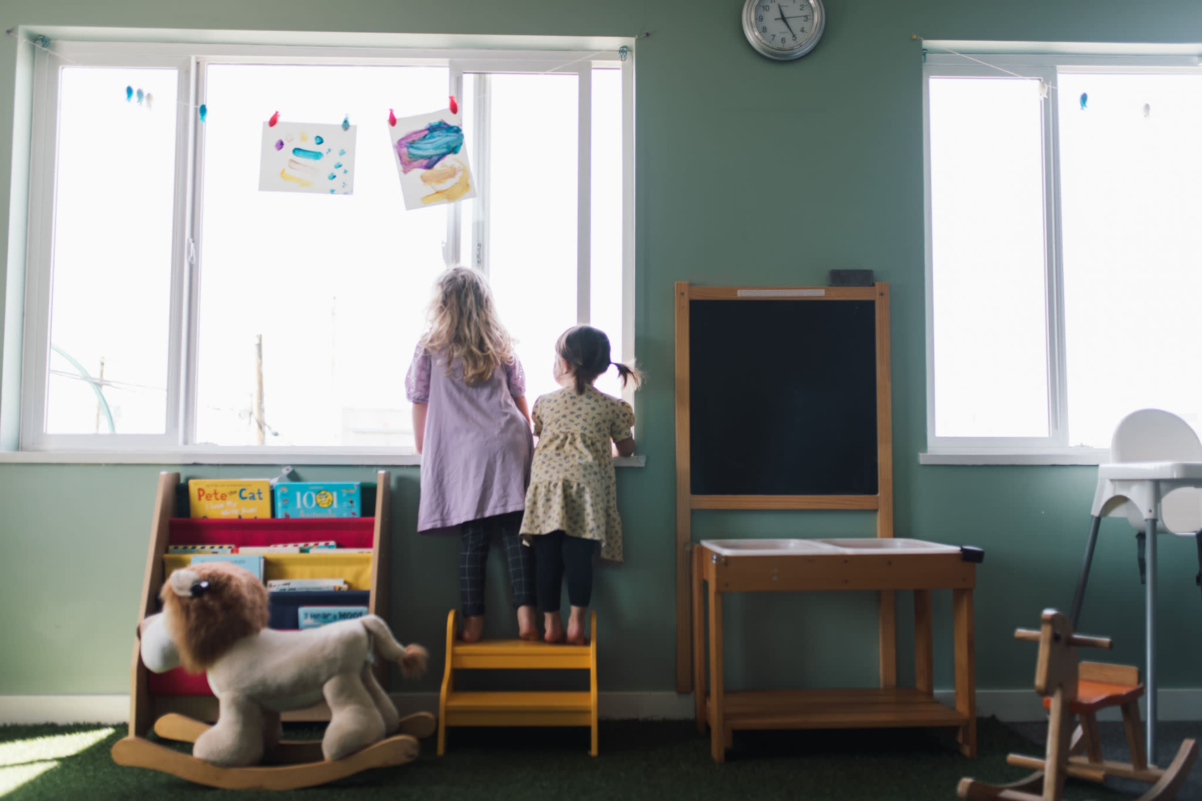 Two children stand by a window in a classroom, looking outside, with a stuffed lion and various toys nearby.
