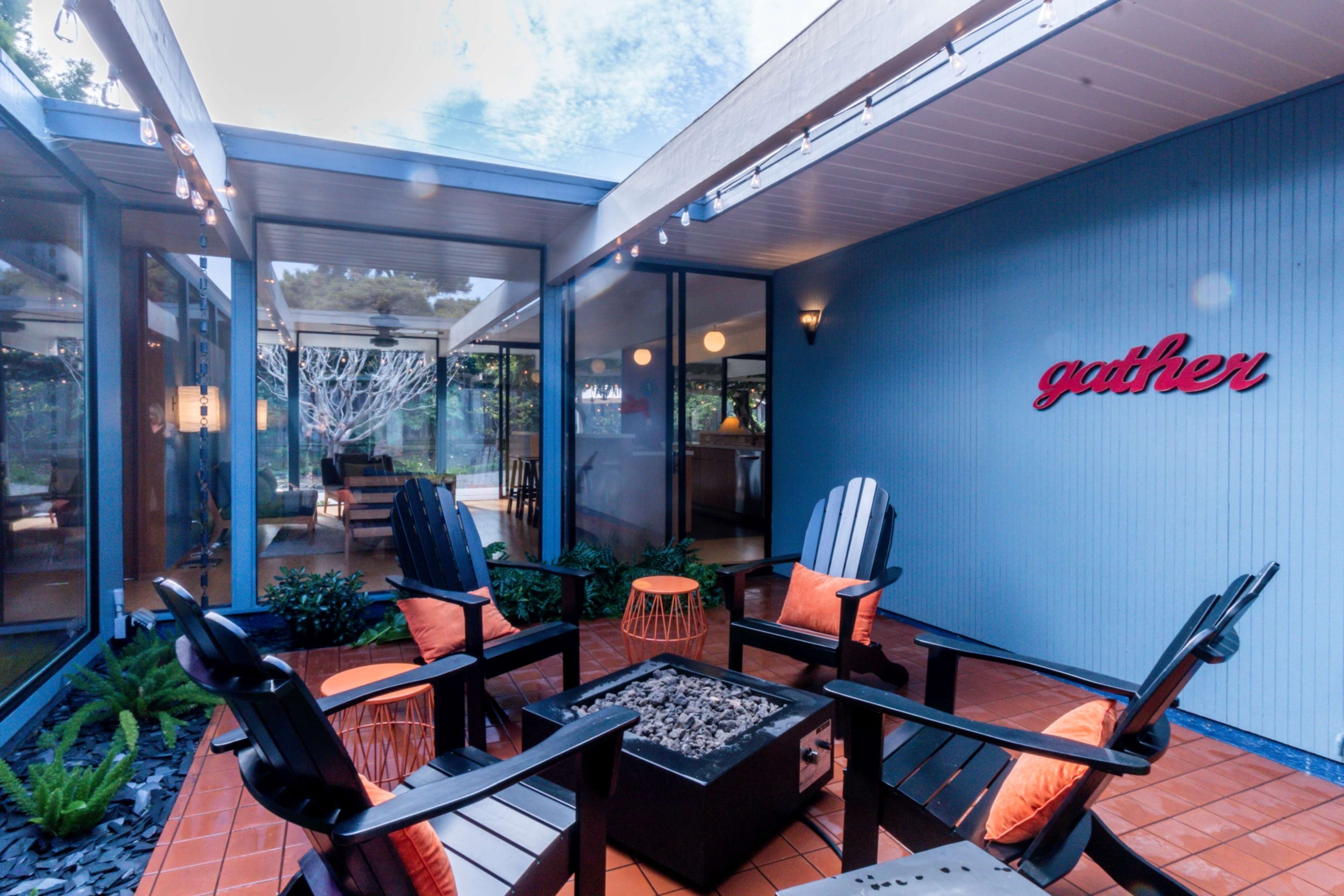 The image shows a patio area with four black chairs arranged around a fire pit, surrounded by glass walls and featuring a wall with the word "gather" in red.