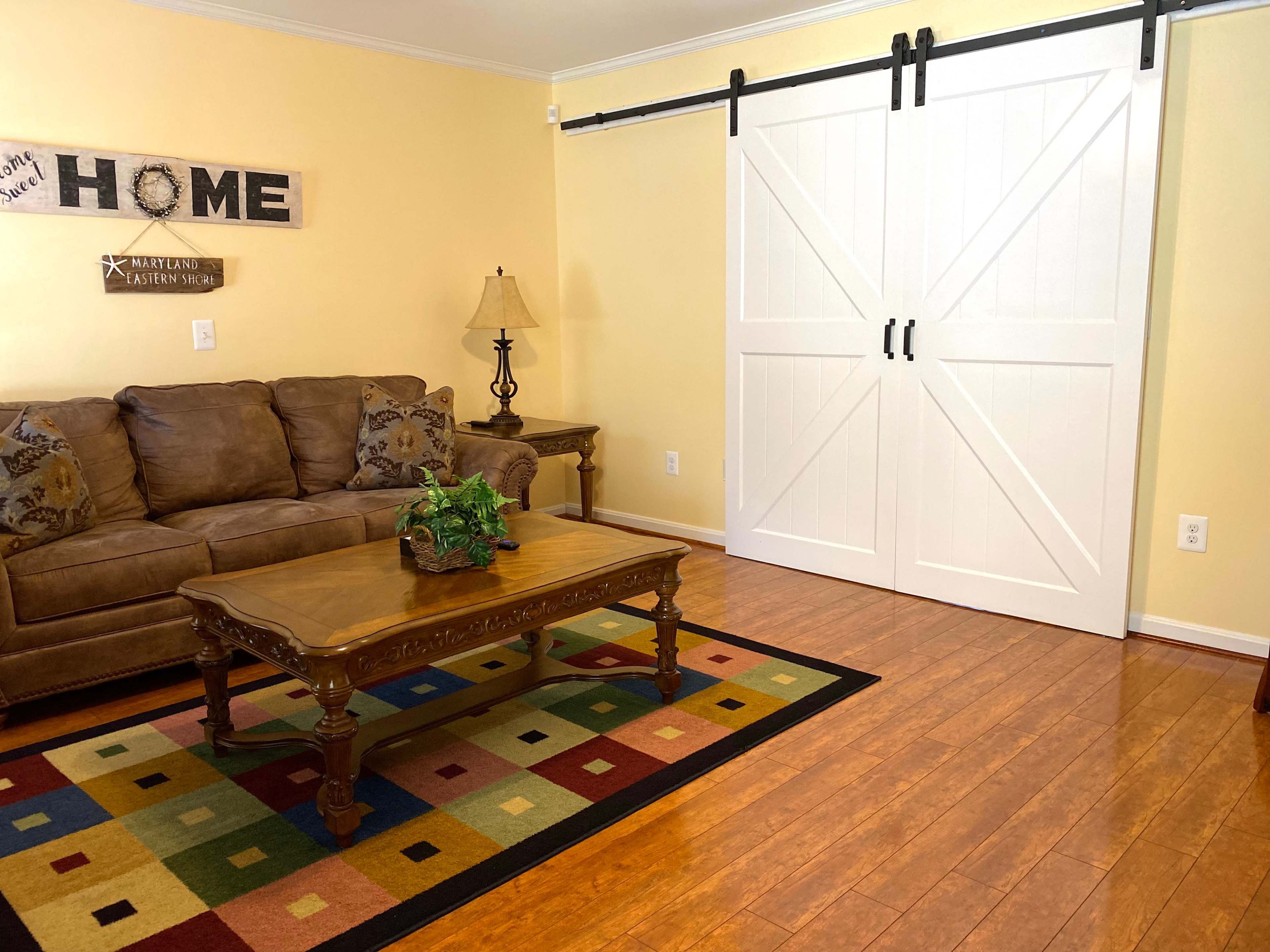 The image shows a cozy living room featuring a brown sofa, a wooden coffee table, a decorative lamp, and a large sliding barn door against a yellow wall.