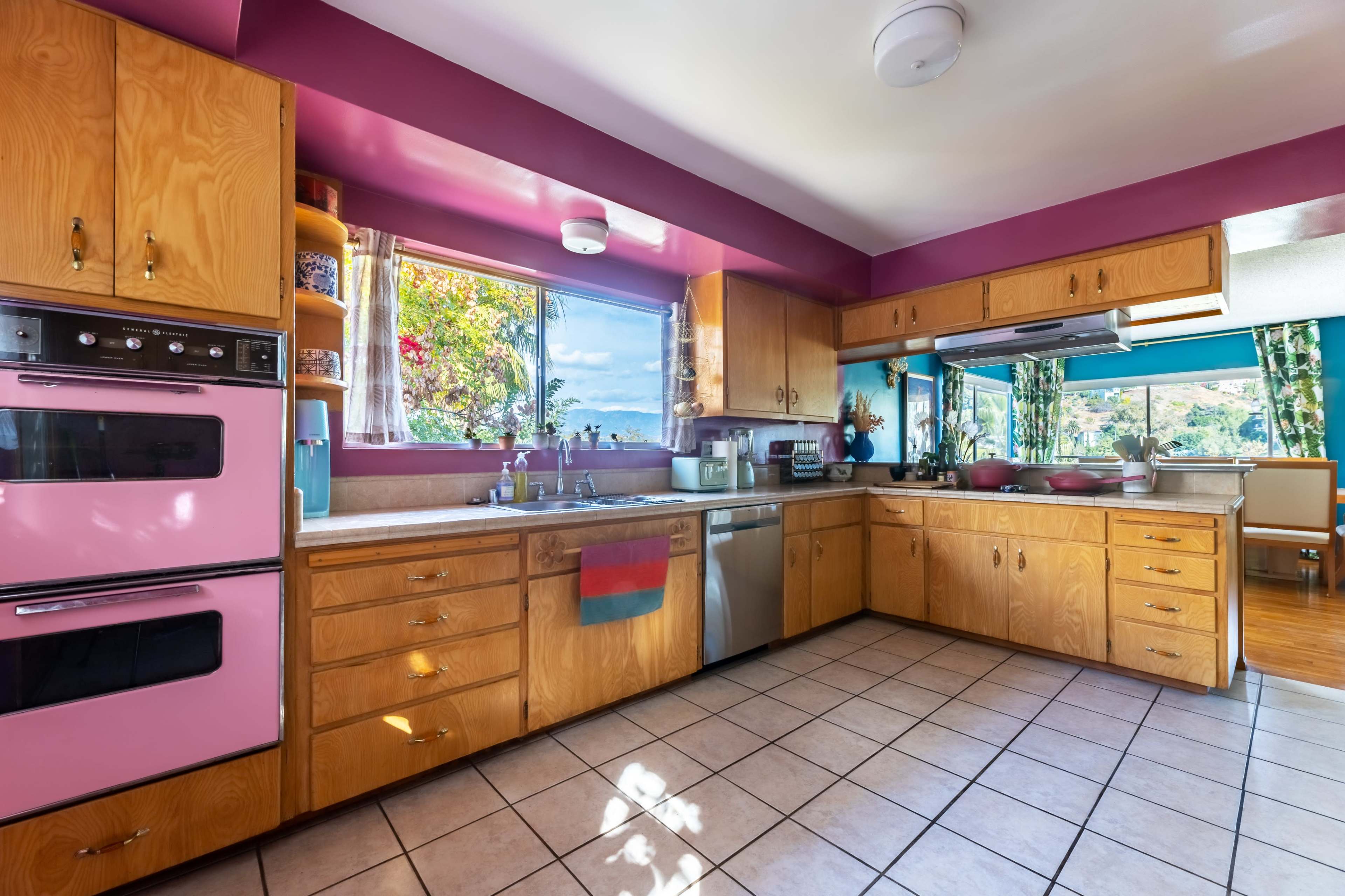 The image shows a bright kitchen with wooden cabinetry, pink accents, and large windows providing natural light and a view of the outdoors.