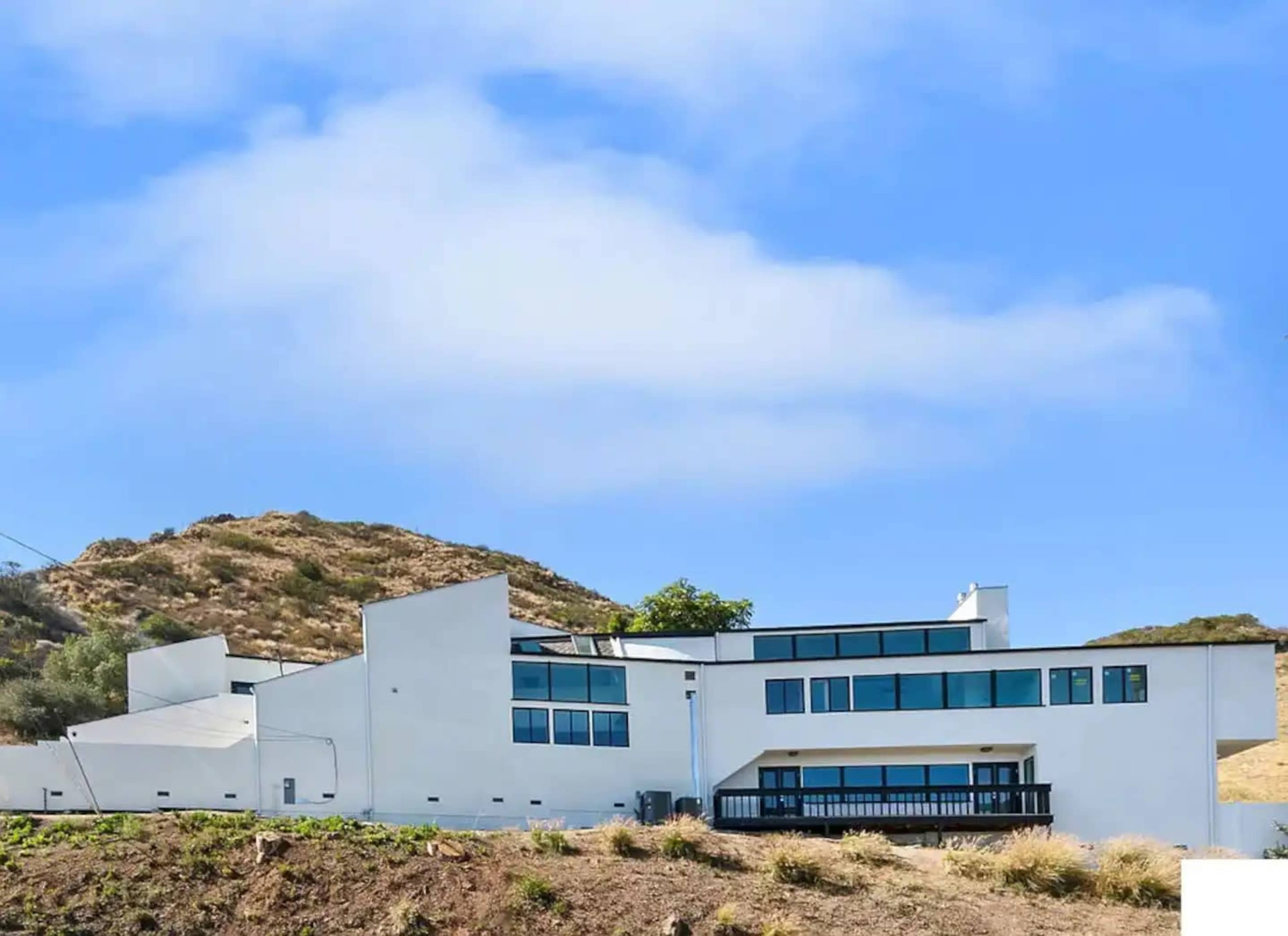 A modern white house with large windows is situated on a hillside under a blue sky.