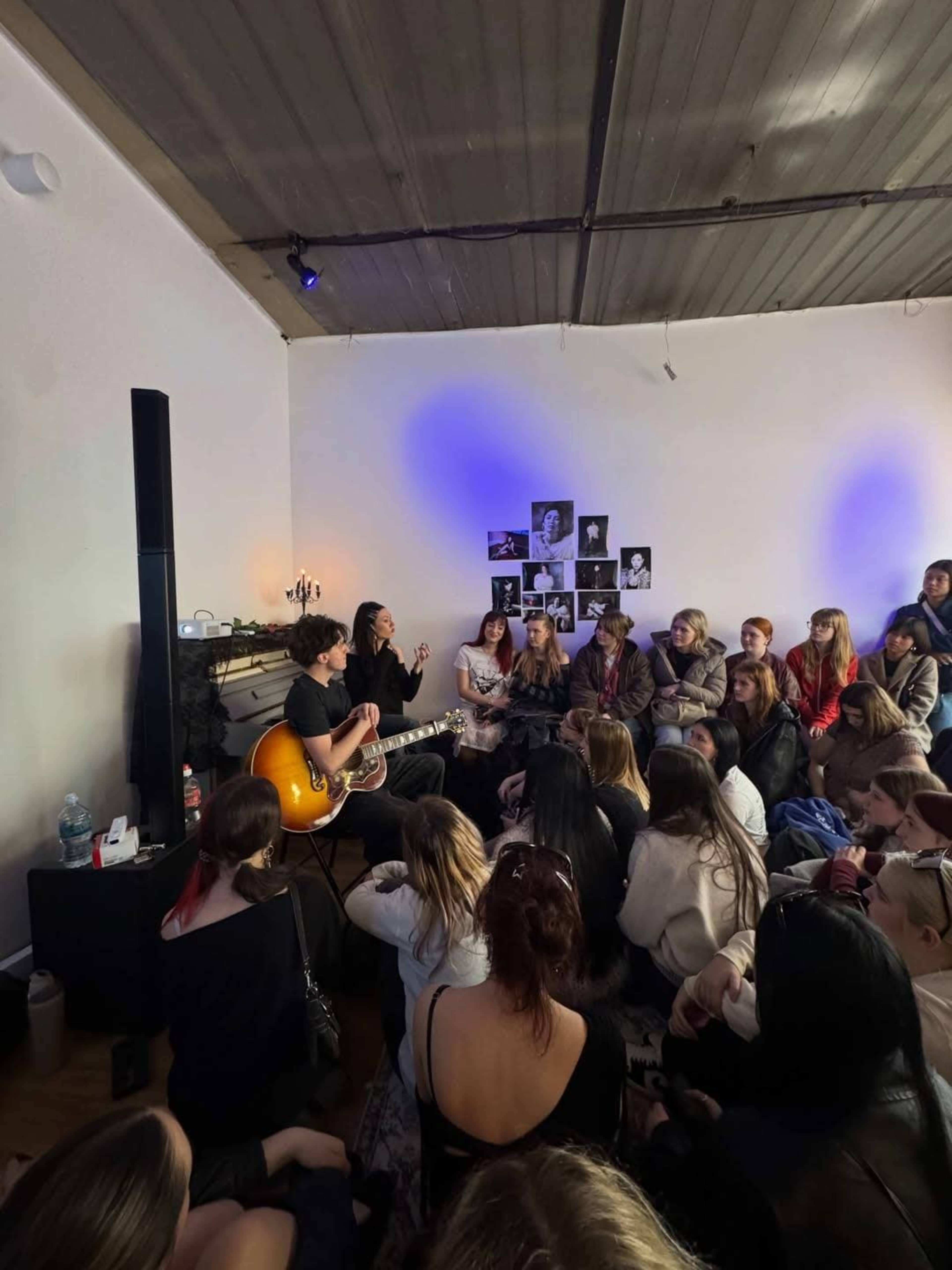 A musician performs an acoustic set in front of an attentive audience seated on the floor in a sparsely decorated room.