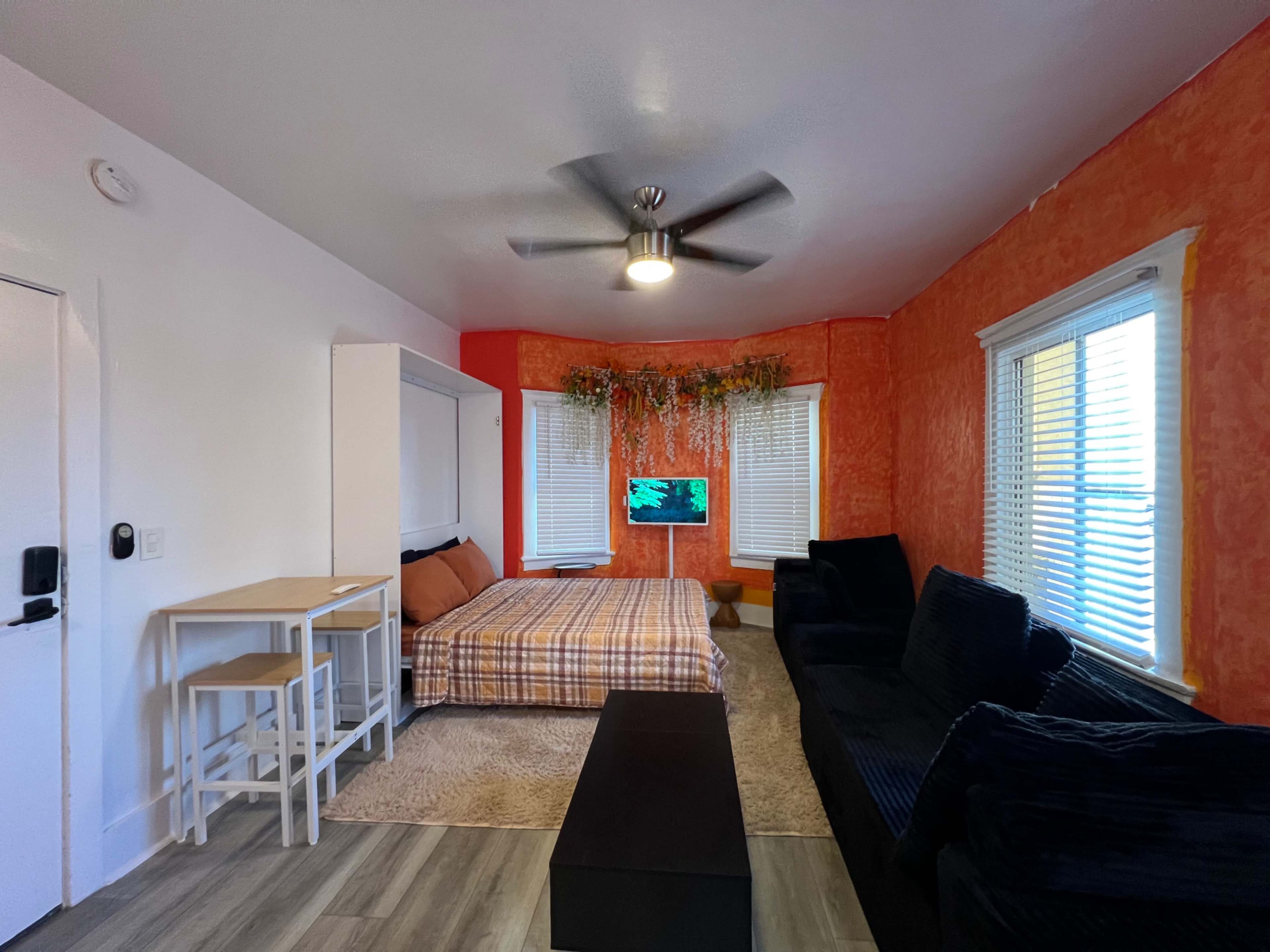 The image shows a living space featuring a foldable bed, a small dining table with chairs, a dark sofa, and a TV mounted on an orange accent wall.