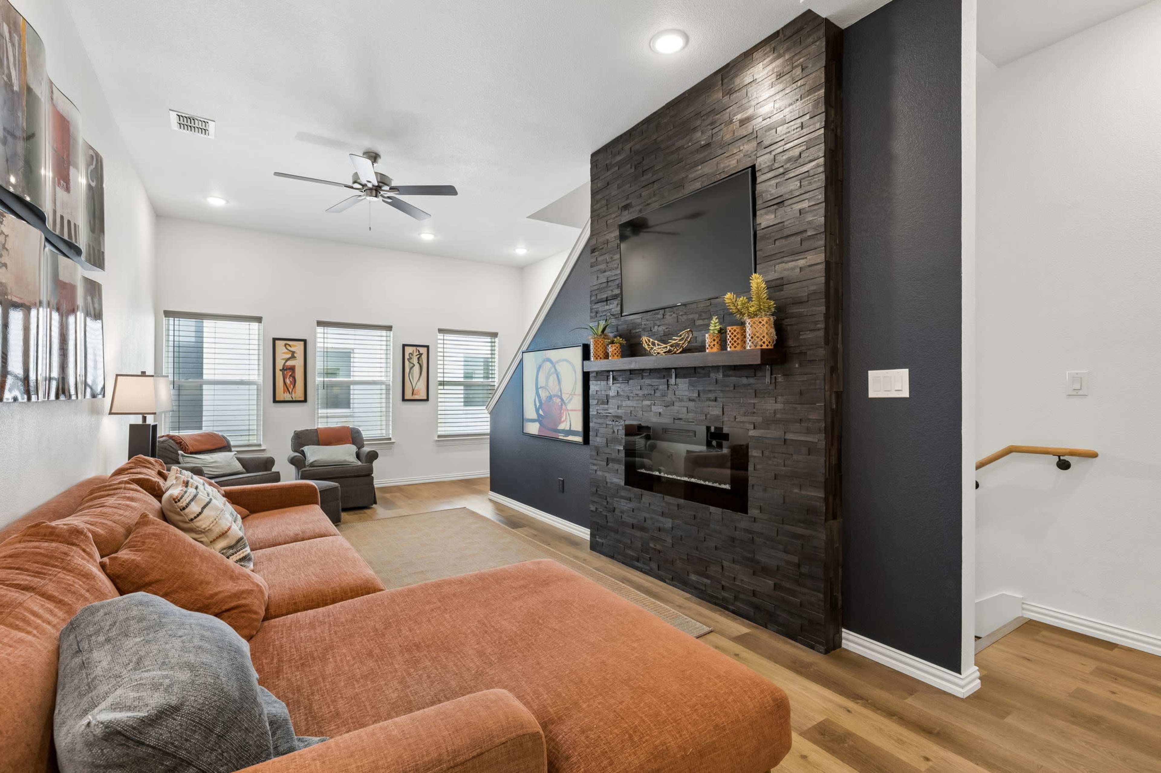 The image shows a modern living room with a sectional sofa, a stone accent wall featuring a flat-screen TV, and large windows allowing natural light.