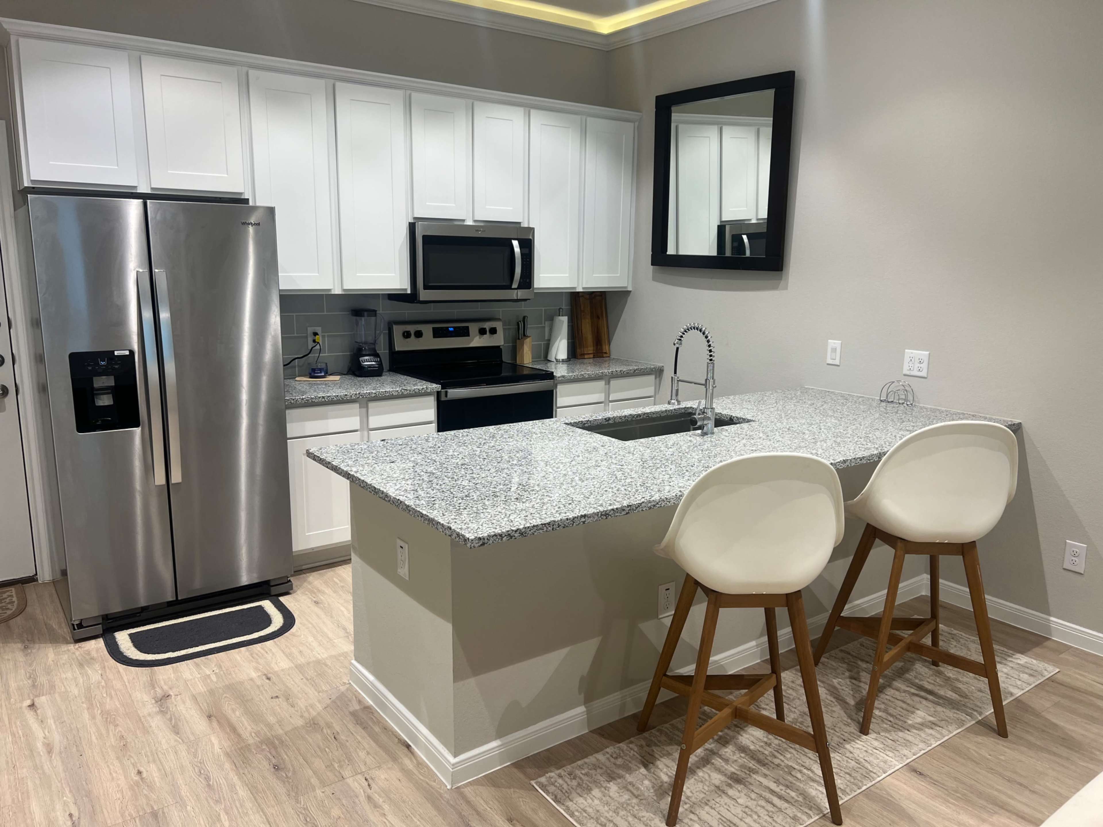 A modern kitchen features white cabinets, stainless steel appliances, a granite countertop island with seating, and a large mirror on the wall.