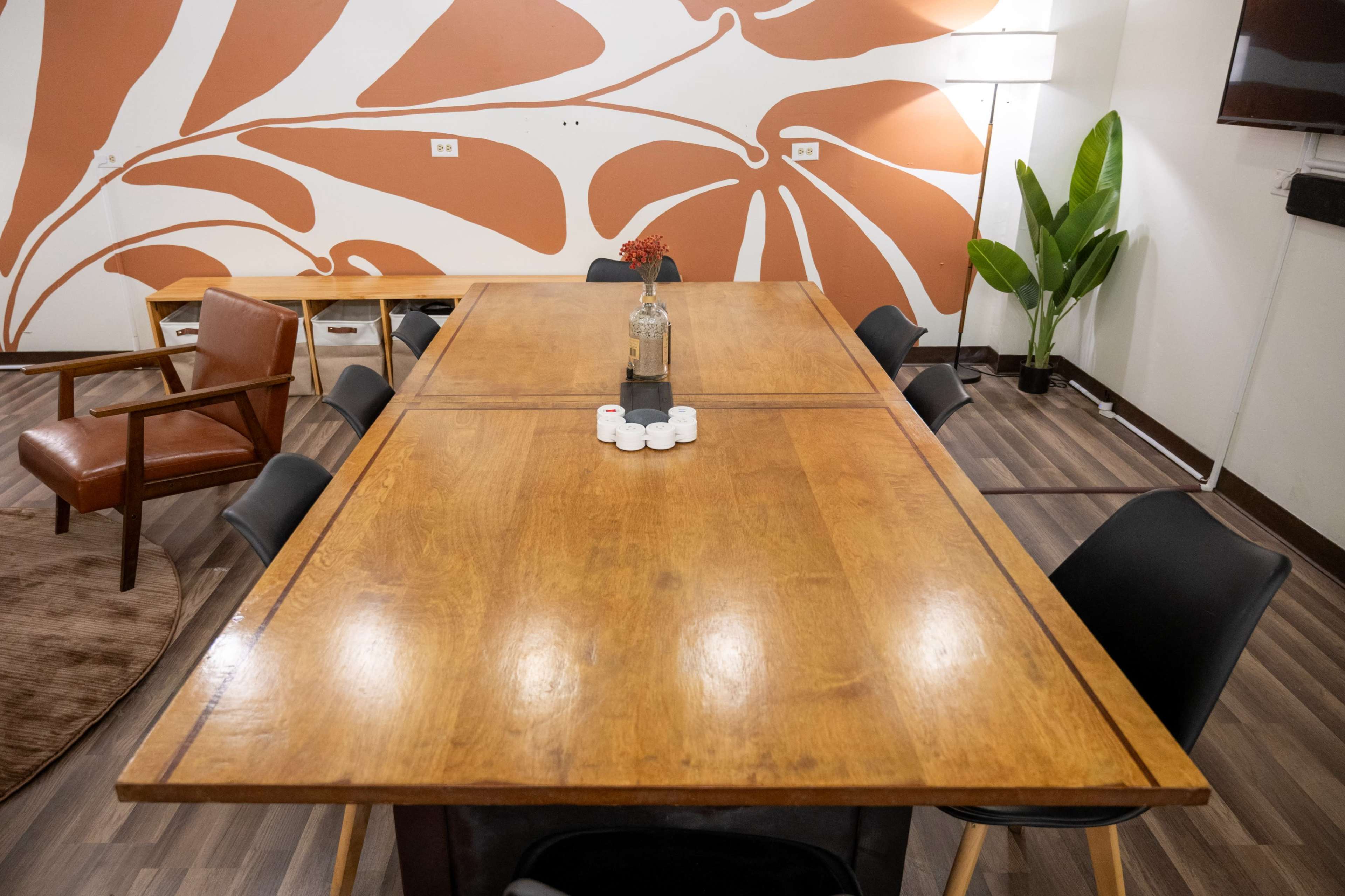 A large wooden table is set up in a well-lit room, surrounded by black chairs, with a decorative plant in the corner and a wall featuring a leafy orange mural.