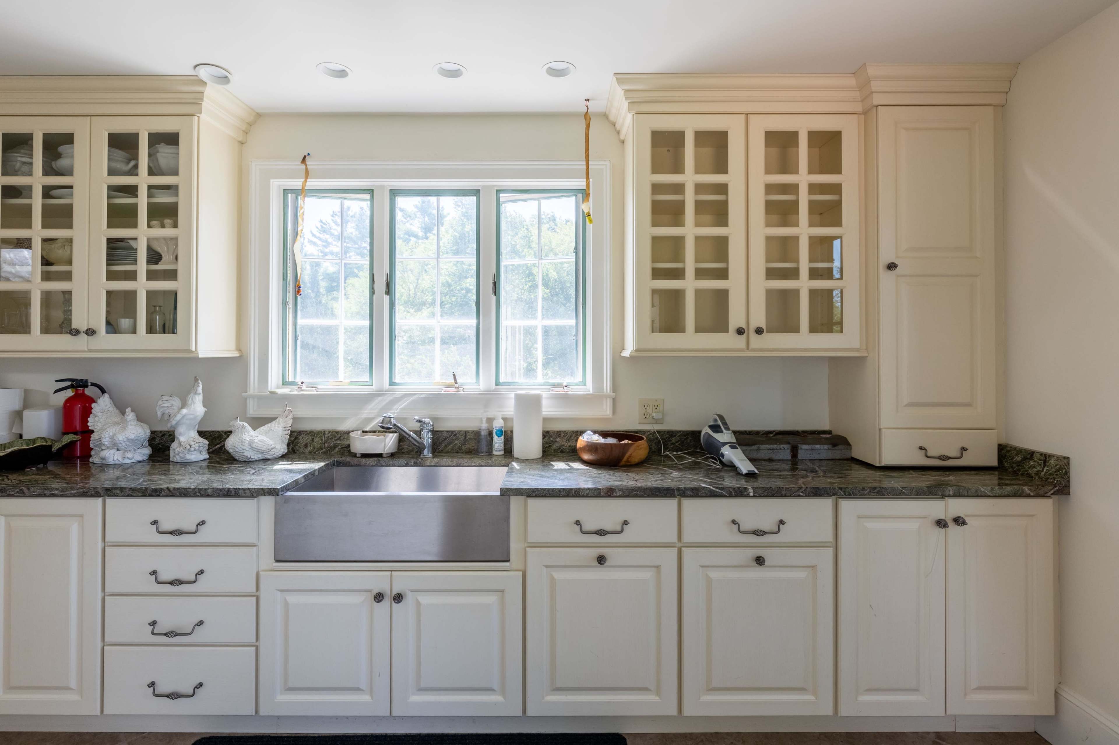 A kitchen features a stainless steel sink beneath a large window, with glass-front cabinets and dark granite countertops.
