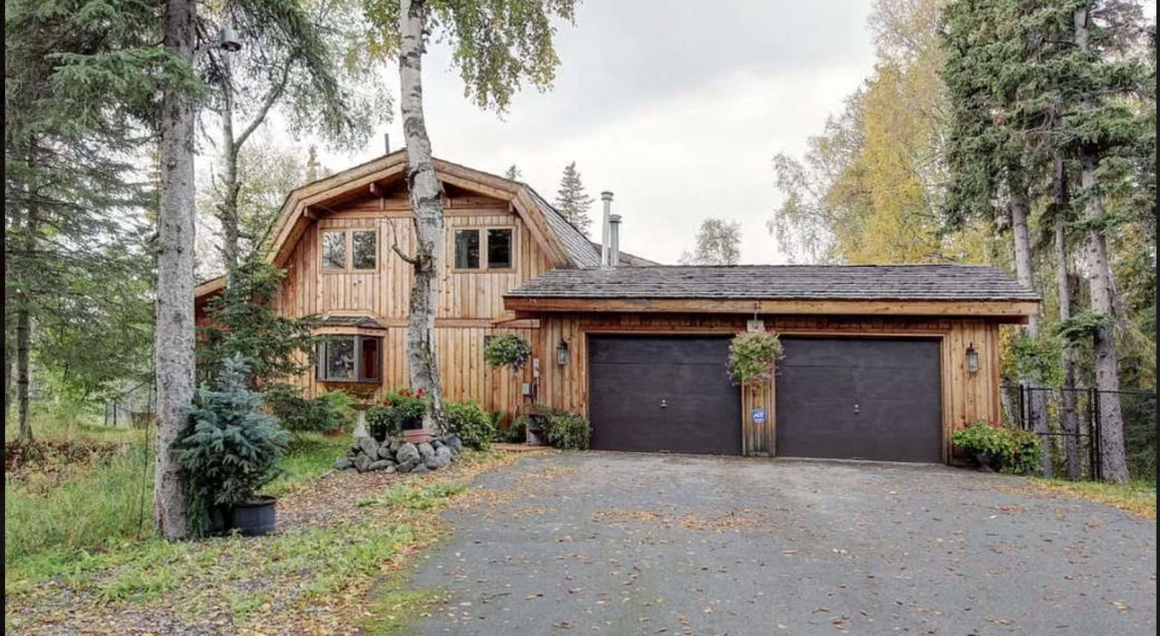A wooden house with a steep roof and a double garage is surrounded by trees and a gravel driveway.