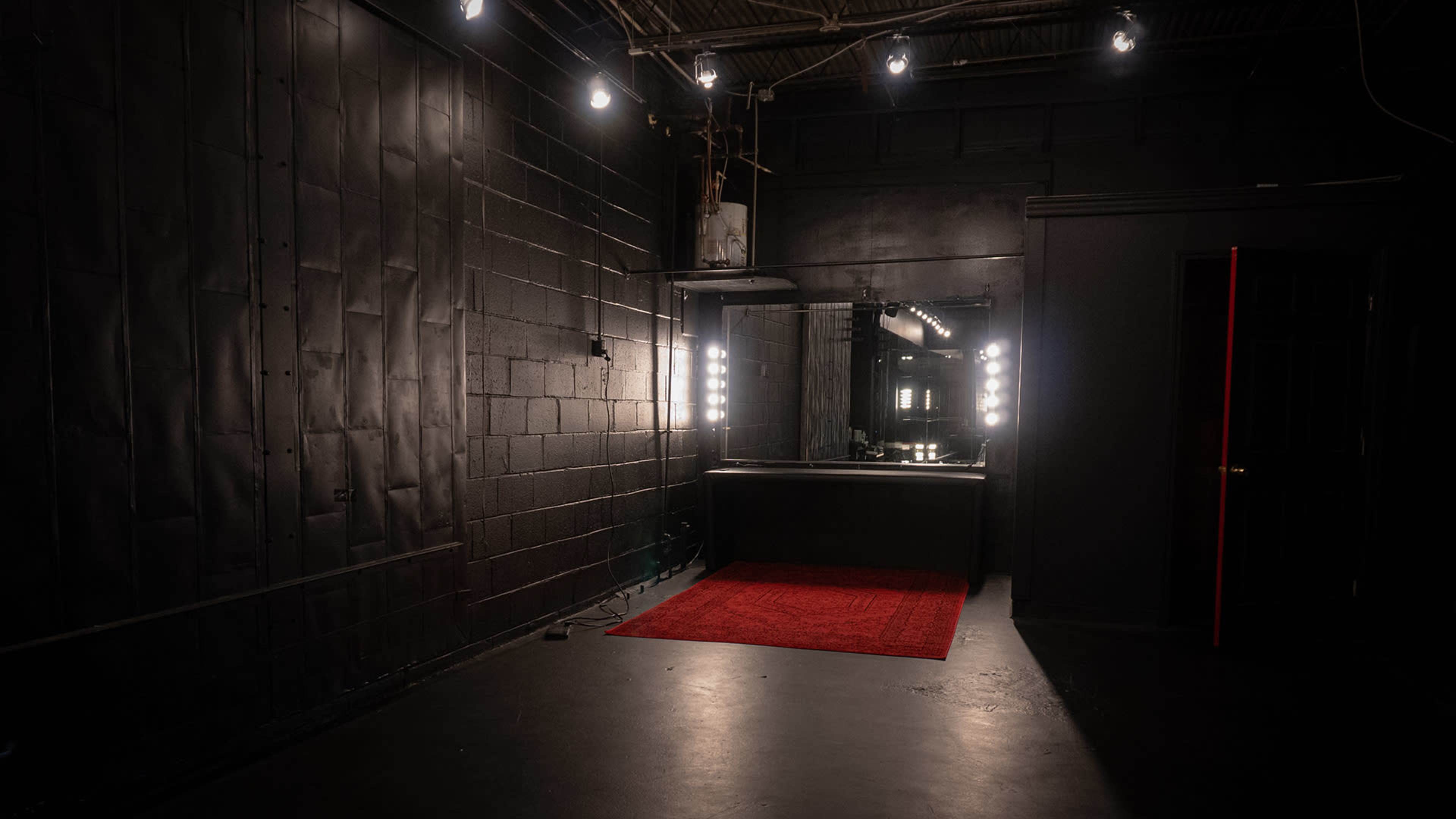 The image shows a dimly lit room with a large mirror and bright lights above a black counter, accompanied by a red carpet on the floor.