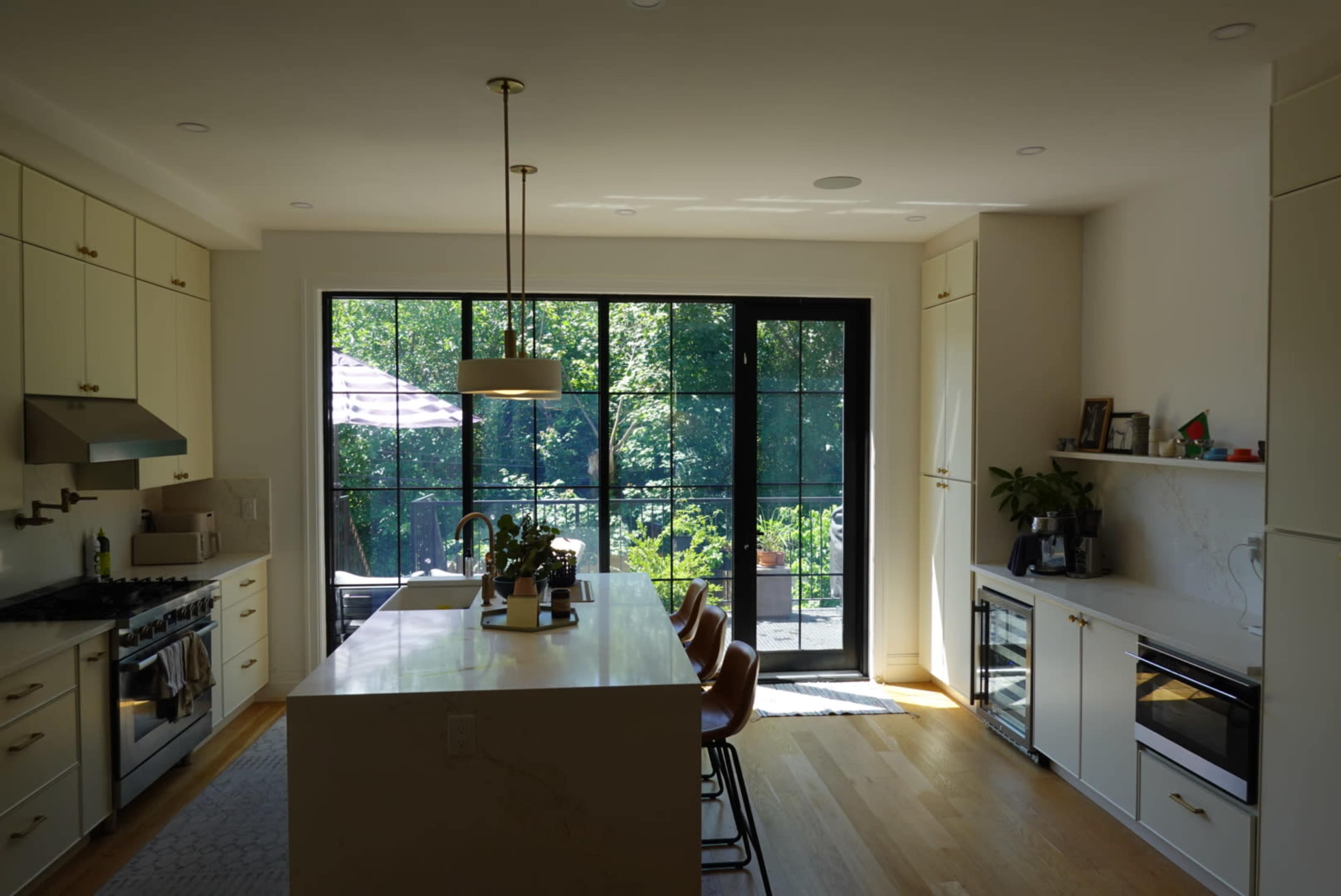 A modern kitchen features a large island with bar seating, stainless steel appliances, and large windows that provide natural light and a view of the outdoors.