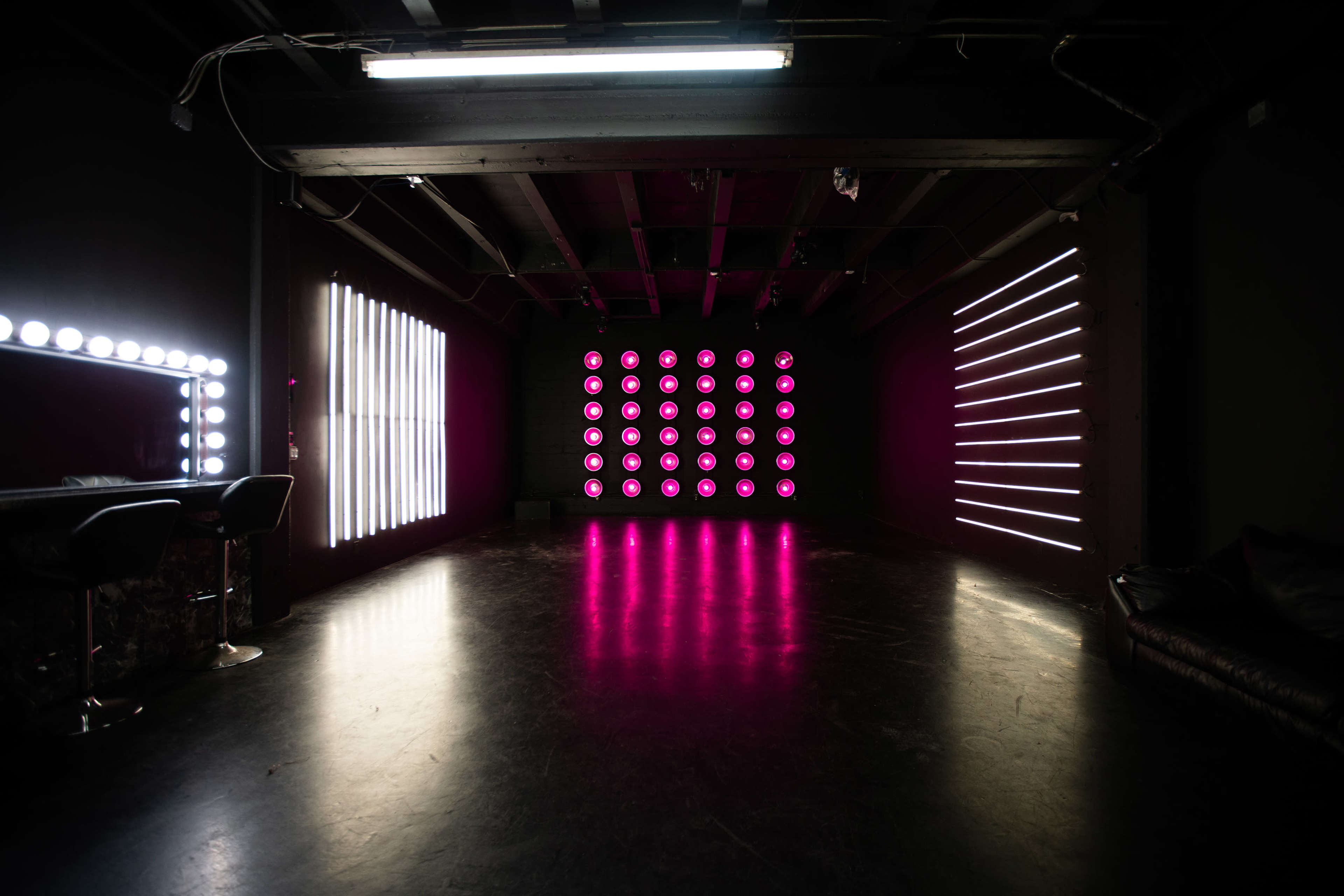 The image shows a dimly lit room featuring a bar area with two stools, multiple light installations on the walls, and a dark floor.