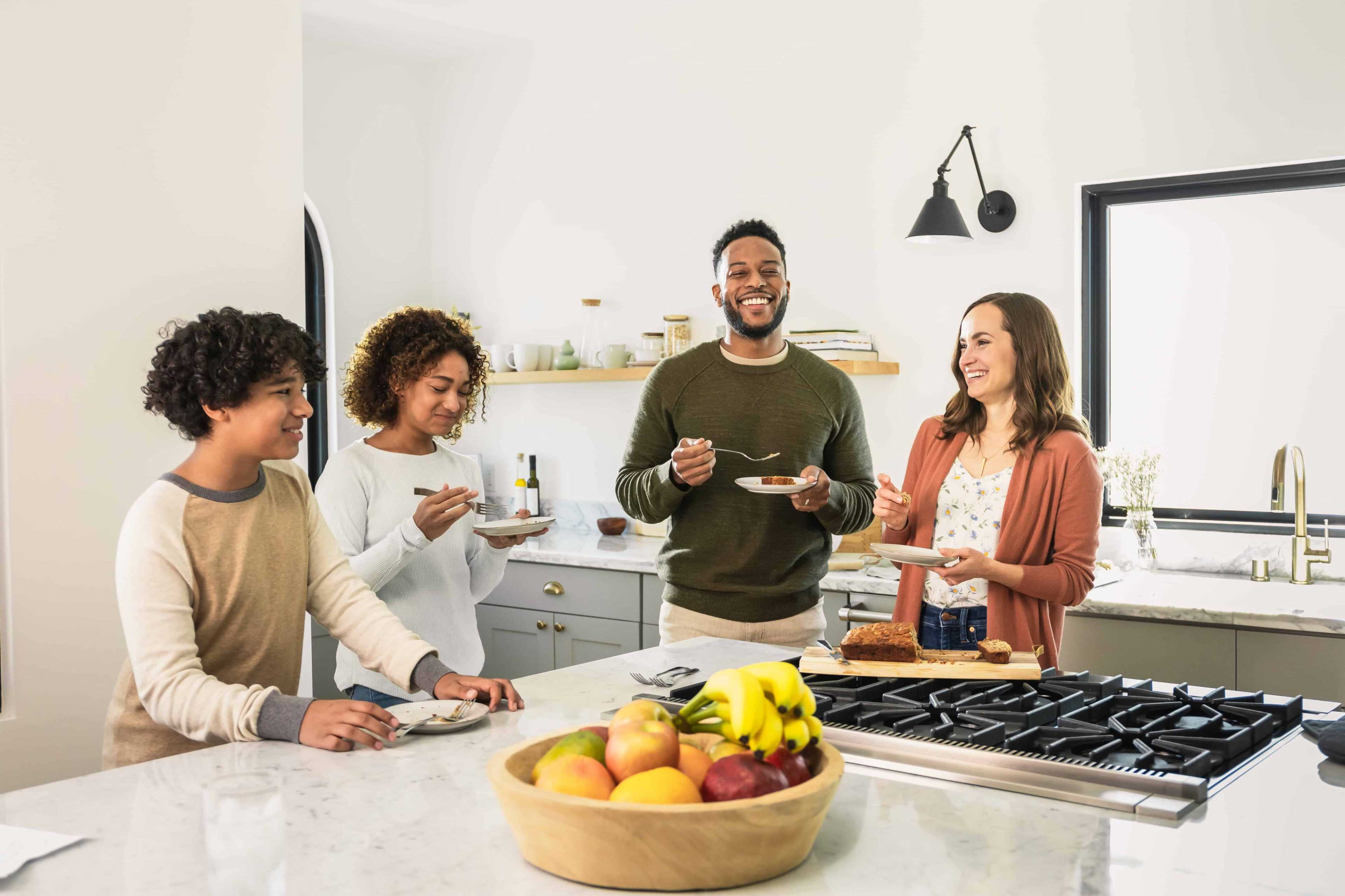 A group of four people enjoys a meal together in a bright kitchen, with a fruit bowl and a stove visible in the background.