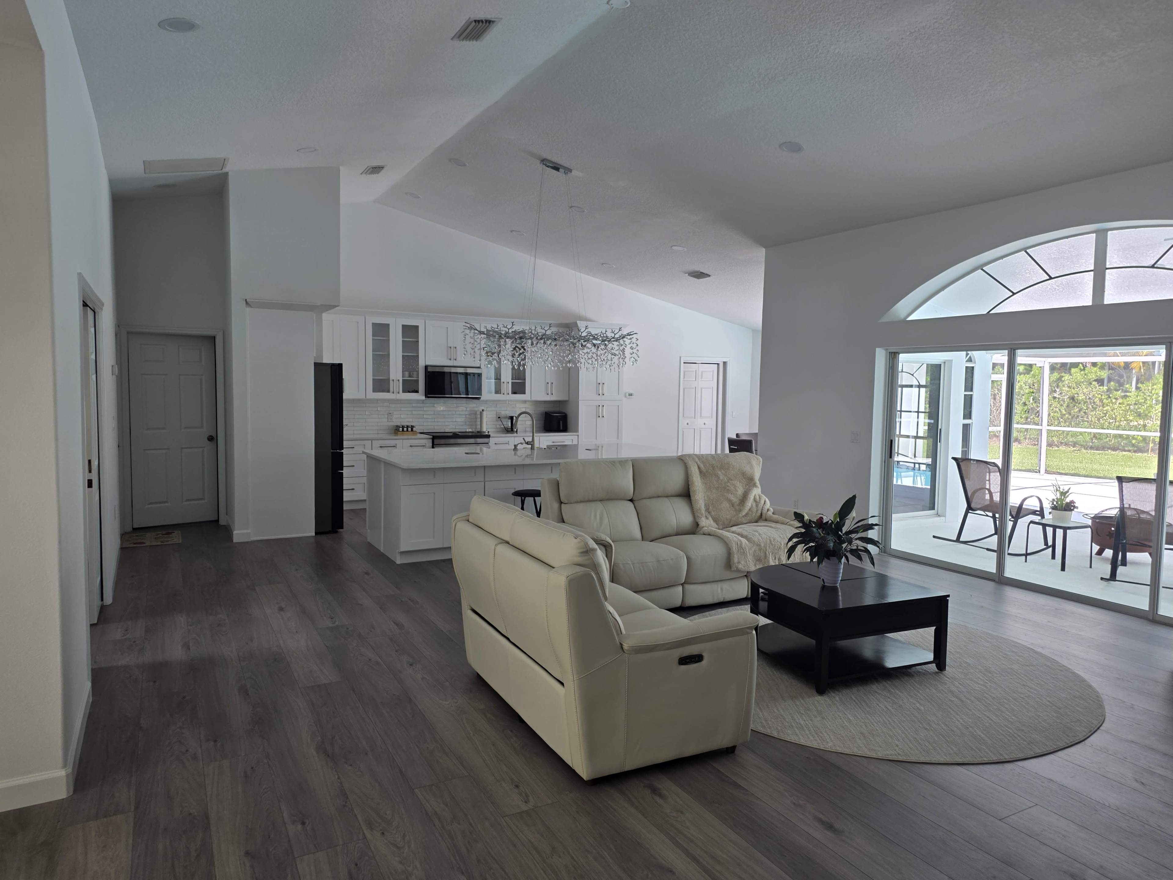The image shows a spacious, bright living room with a white leather sofa, a black coffee table, and a view of a pool area through large windows.
