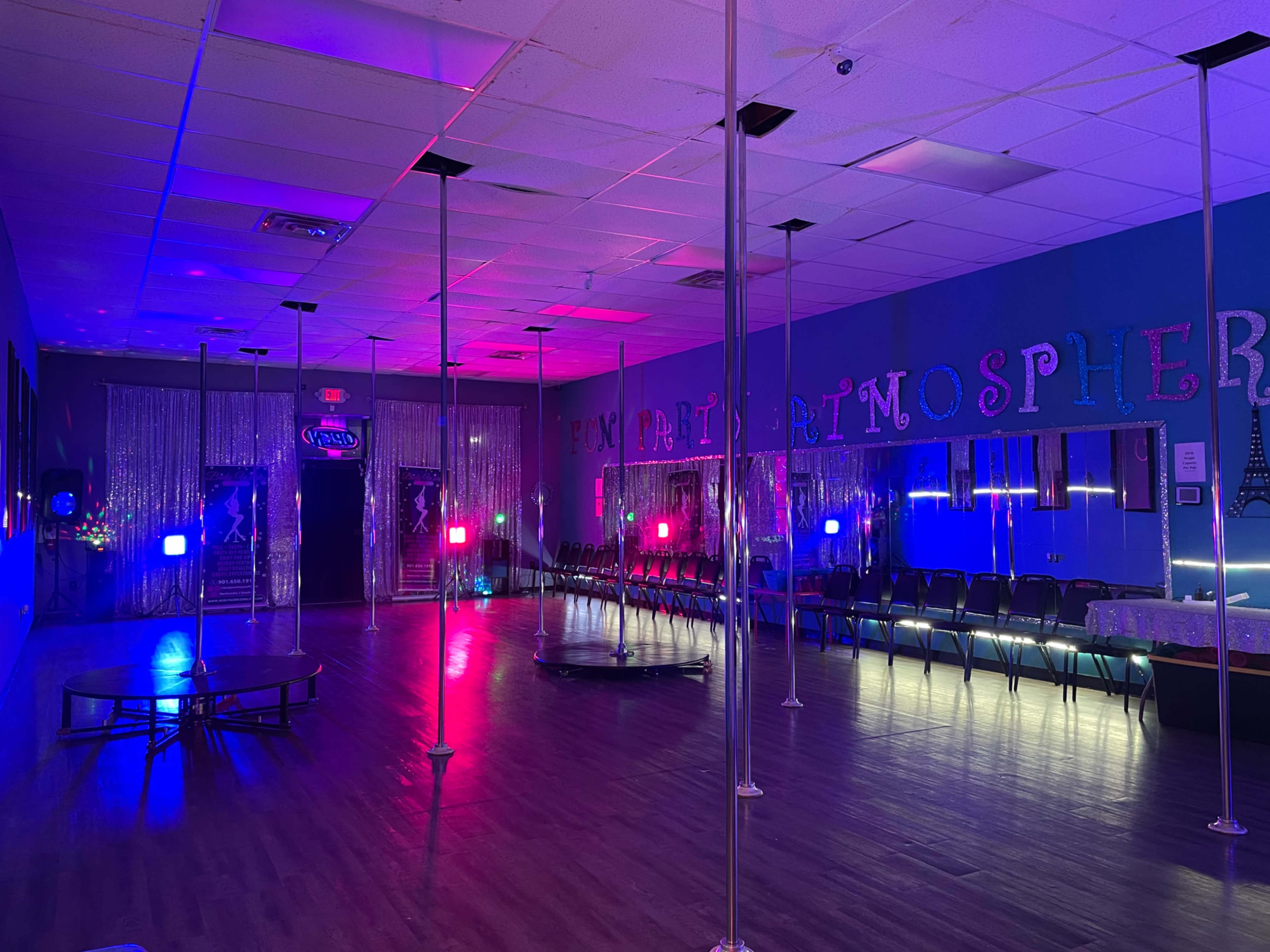 A dance studio featuring several poles, colorful lights, and rows of black chairs along the walls.